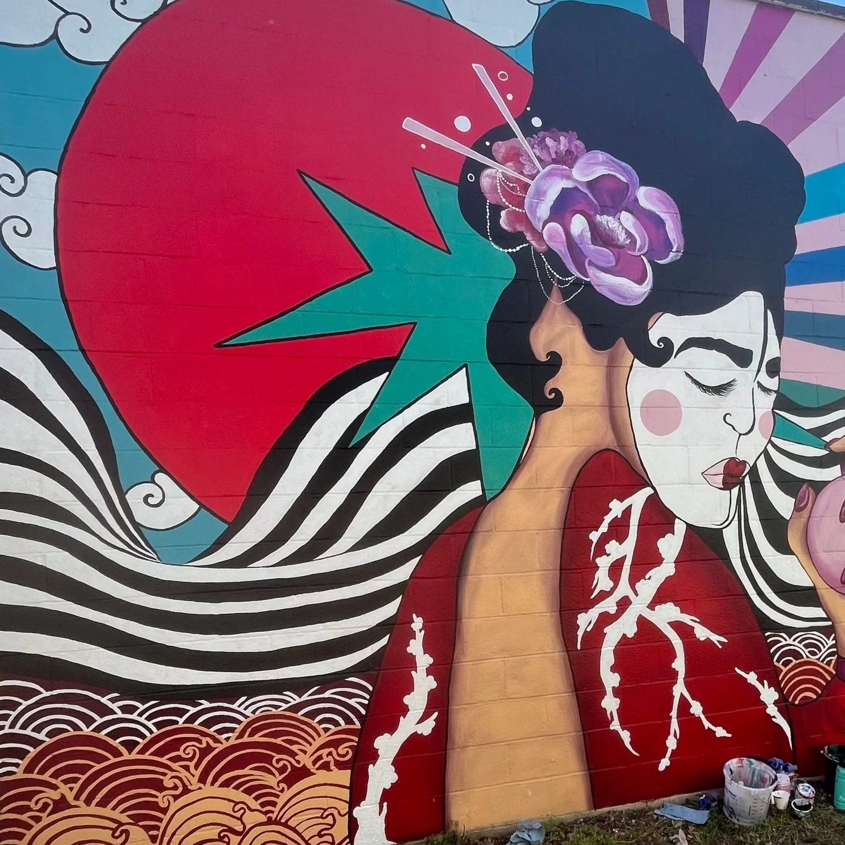 Colorful mural of a woman with black hair, wearing a red kimono with white tree branches, holding a pink fan, with a large pink sun, black and white striped waves, clouds, and a purple flower in her hair.