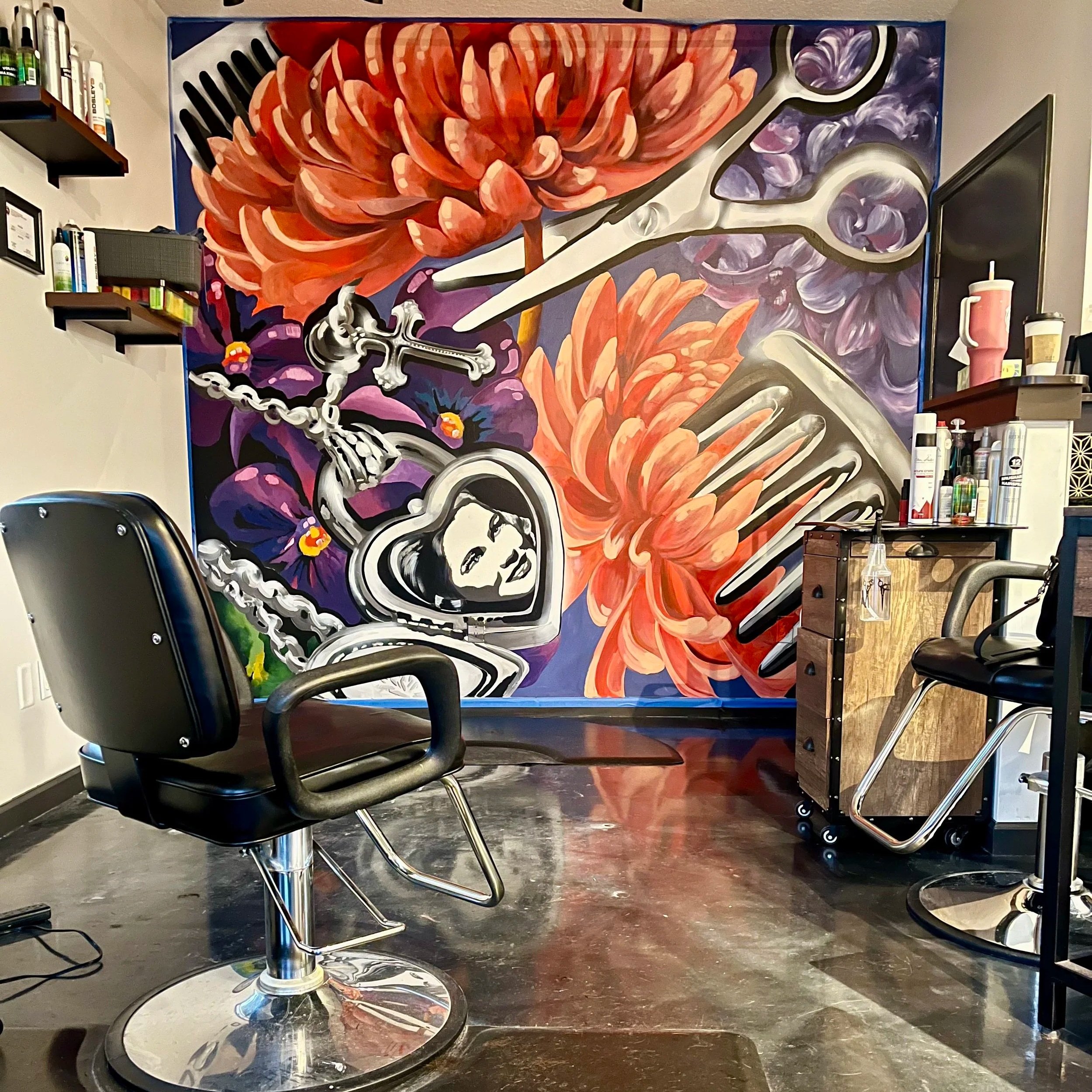 Interior view of a hair salon with a large mural wall featuring vibrant flowers, scissors, a comb, and a woman's face in a heart-shaped frame, along with salon chairs and styling tools.