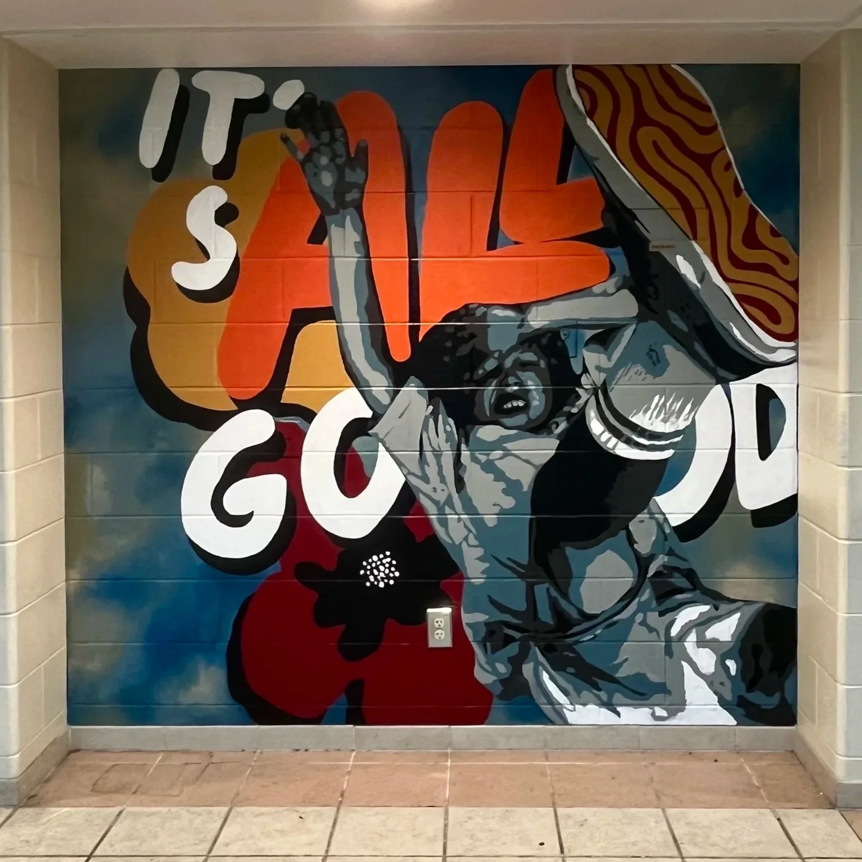A colorful mural depicting a person holding a large pizza. The person appears to be excited or cheering, with background text reading "IT'S ALL GOOD." The mural is painted on a tiled wall with bright colors including red, orange, yellow, blue, and black.