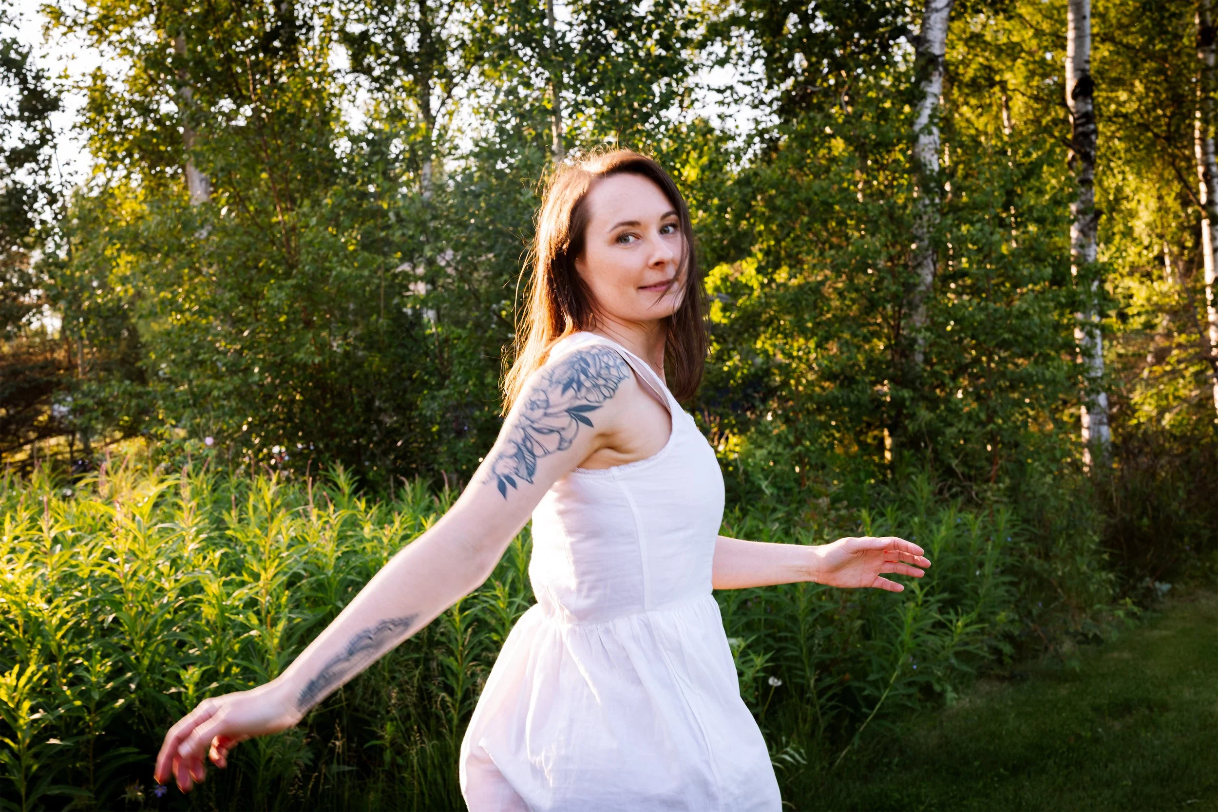 A woman wearing a white sleeveless dress looking back at the camera with a smile as she runs through a lush green field.