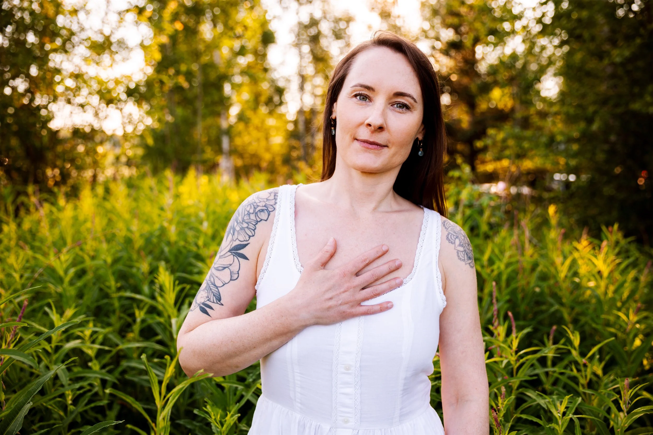 A woman with floral tattoos on her arm standing outdoors in a sunlit field with trees in the background, holding her hand to her chest as she looks gently at the camera.