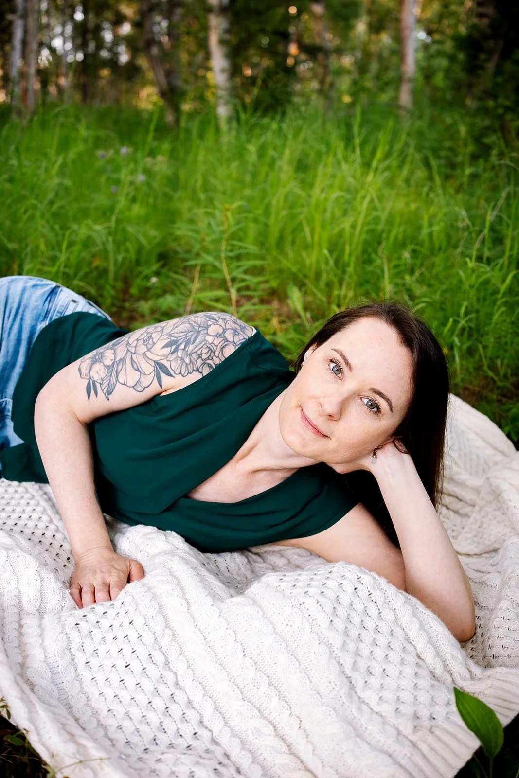A woman with brown hair and a floral tattoo on her arm, lying on her side on a knitted white blanket in a bright green field beneath trees, looks at the camera with a soft and comforting smile.