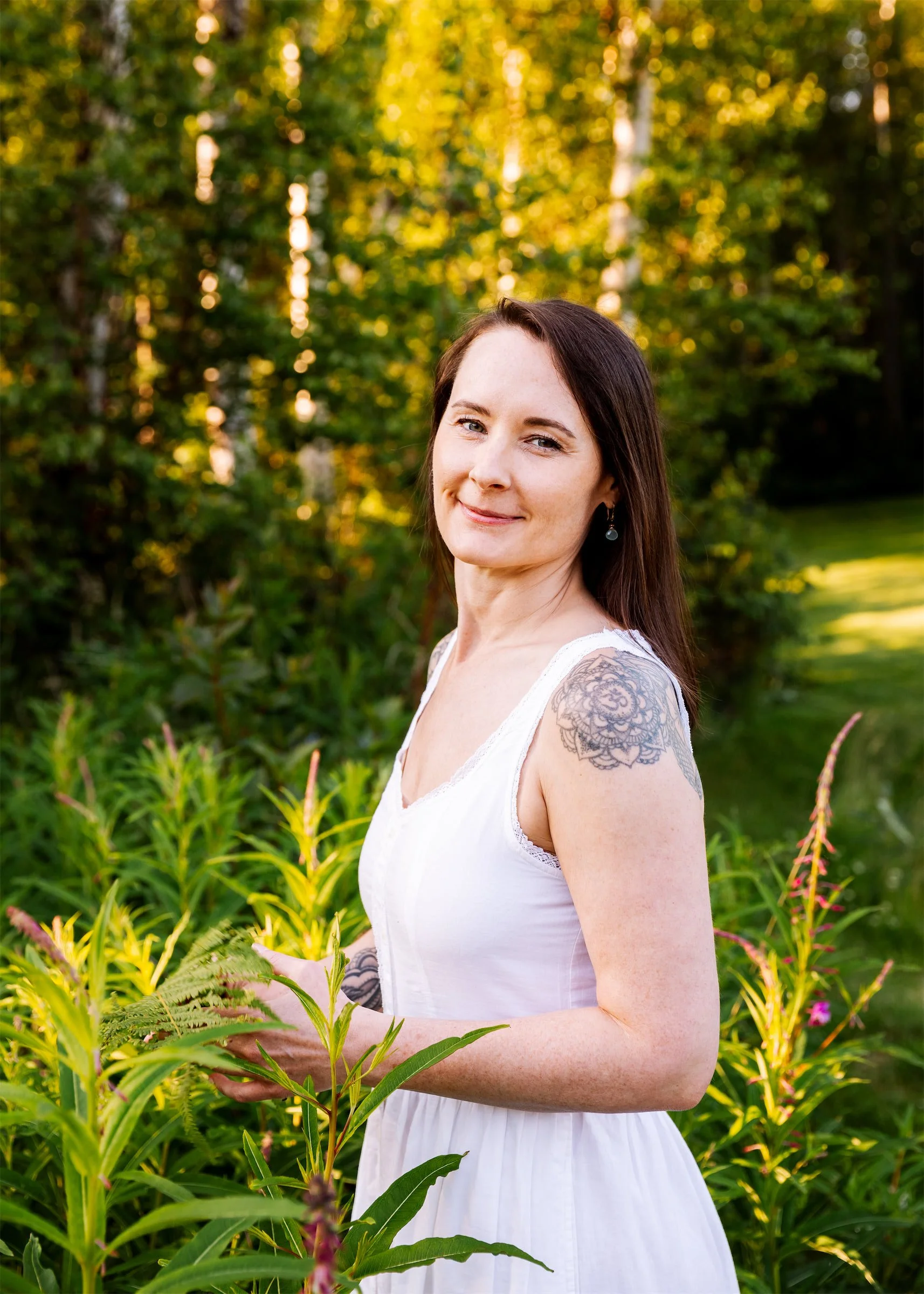 A softly smiling woman with long brown hair, wearing a white dress, standing outdoors in a lush green garden with tall trees in the background.