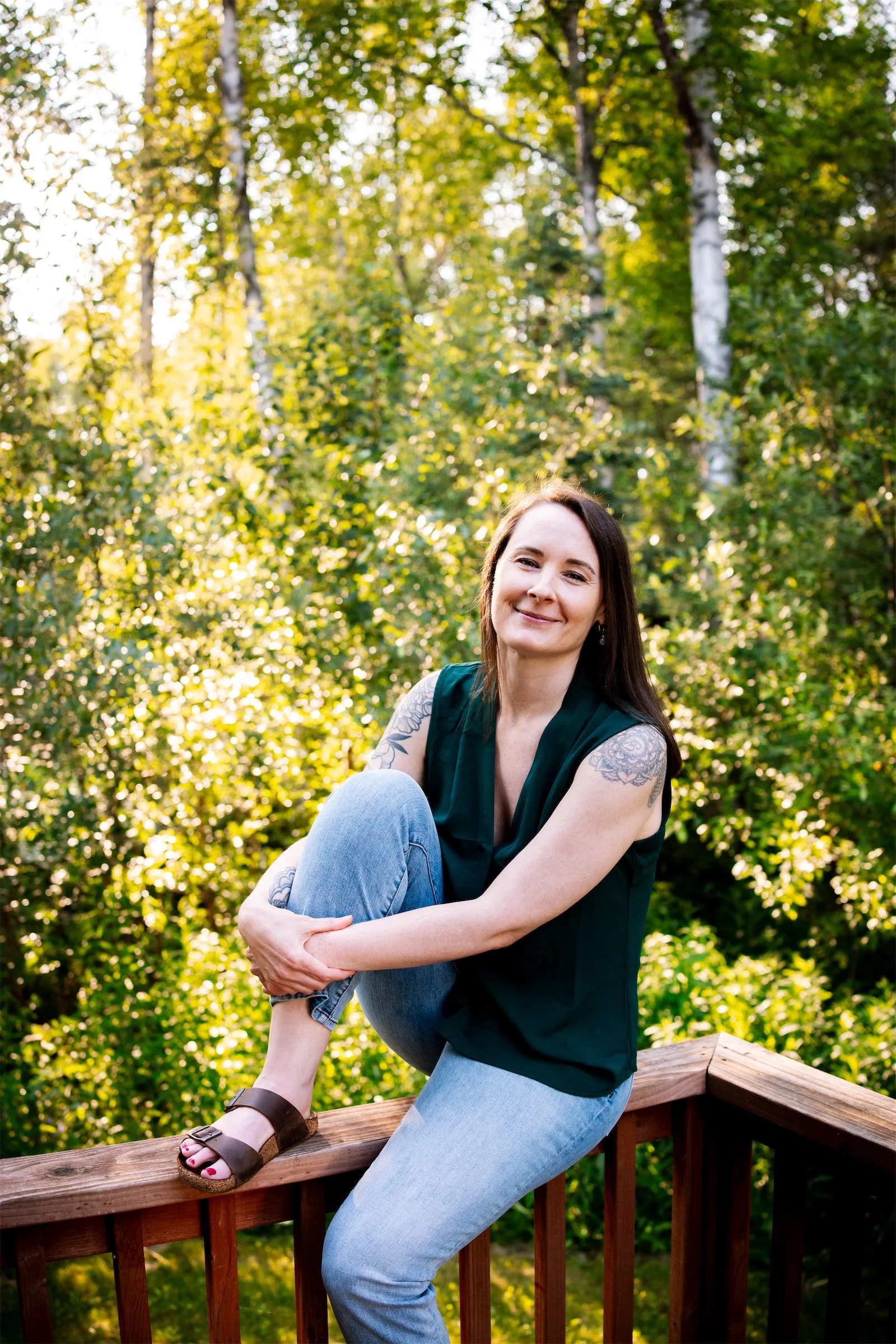 A friendly smiling woman sitting on a wooden deck railing in a forested area with one foot up on the railing, wearing a sleeveless dark green top, light blue jeans, and sandals, with tattoos on her arms.