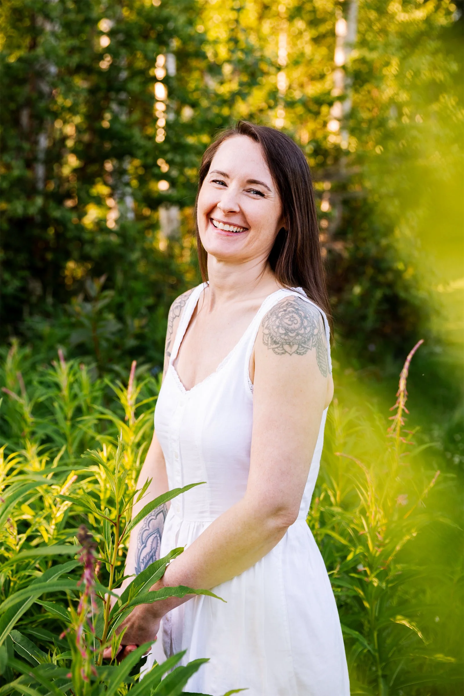 Woman with long dark hair in a white sleeveless dress, standing in a field of waist-high fireweed plants, smiling wide and joyfully at the camera with her head slightly tilted back.