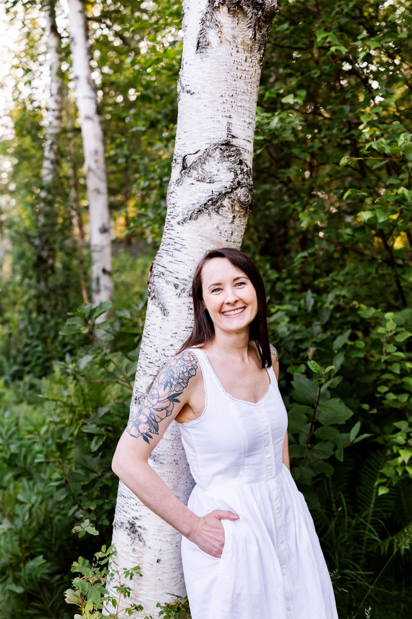 A woman leans against a white birch tree trunk with with her hands in the pockets of her white sleeveless dress, looking at the camera with a welcoming smile.
