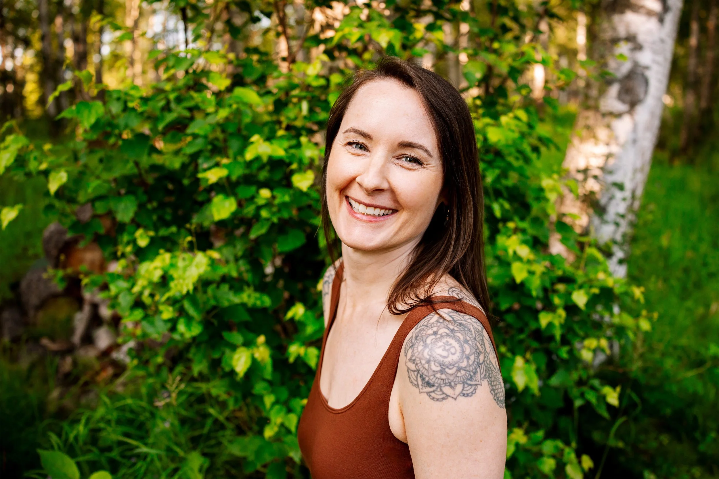 A brightly smiling woman with long brown hair, wearing a brown tank top dress, standing in front of green foliage and trees.