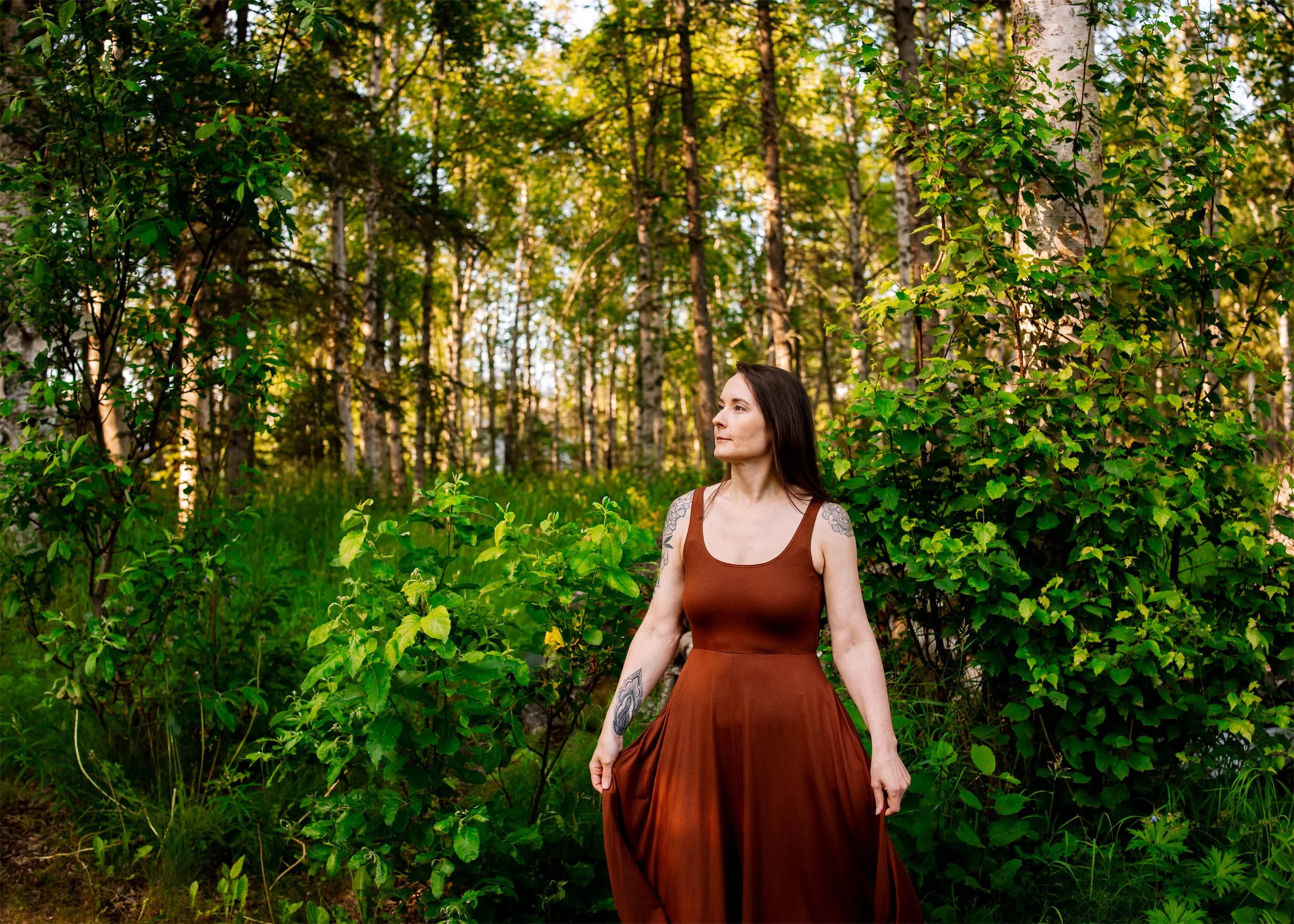A woman with long dark hair, wearing a rust-colored sleeveless dress, stands in a forest with tall trees and green foliage, looking off into the distance.