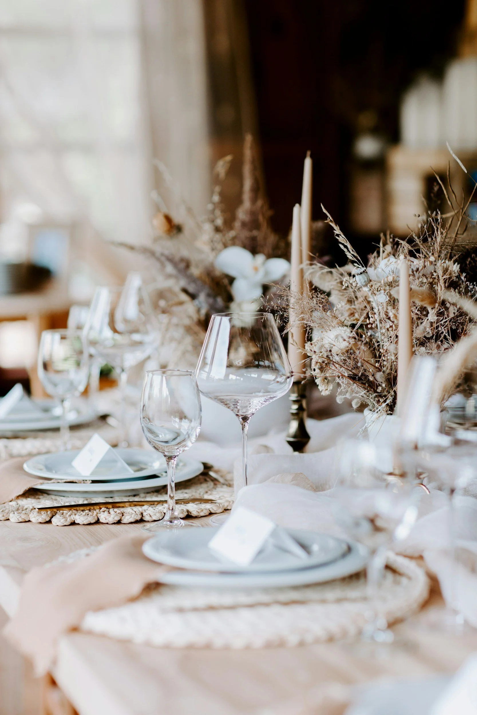 A table setting with clear wine glasses, white plates, and a floral centerpiece with dried flowers and candles, prepared for a formal event or dinner party.
