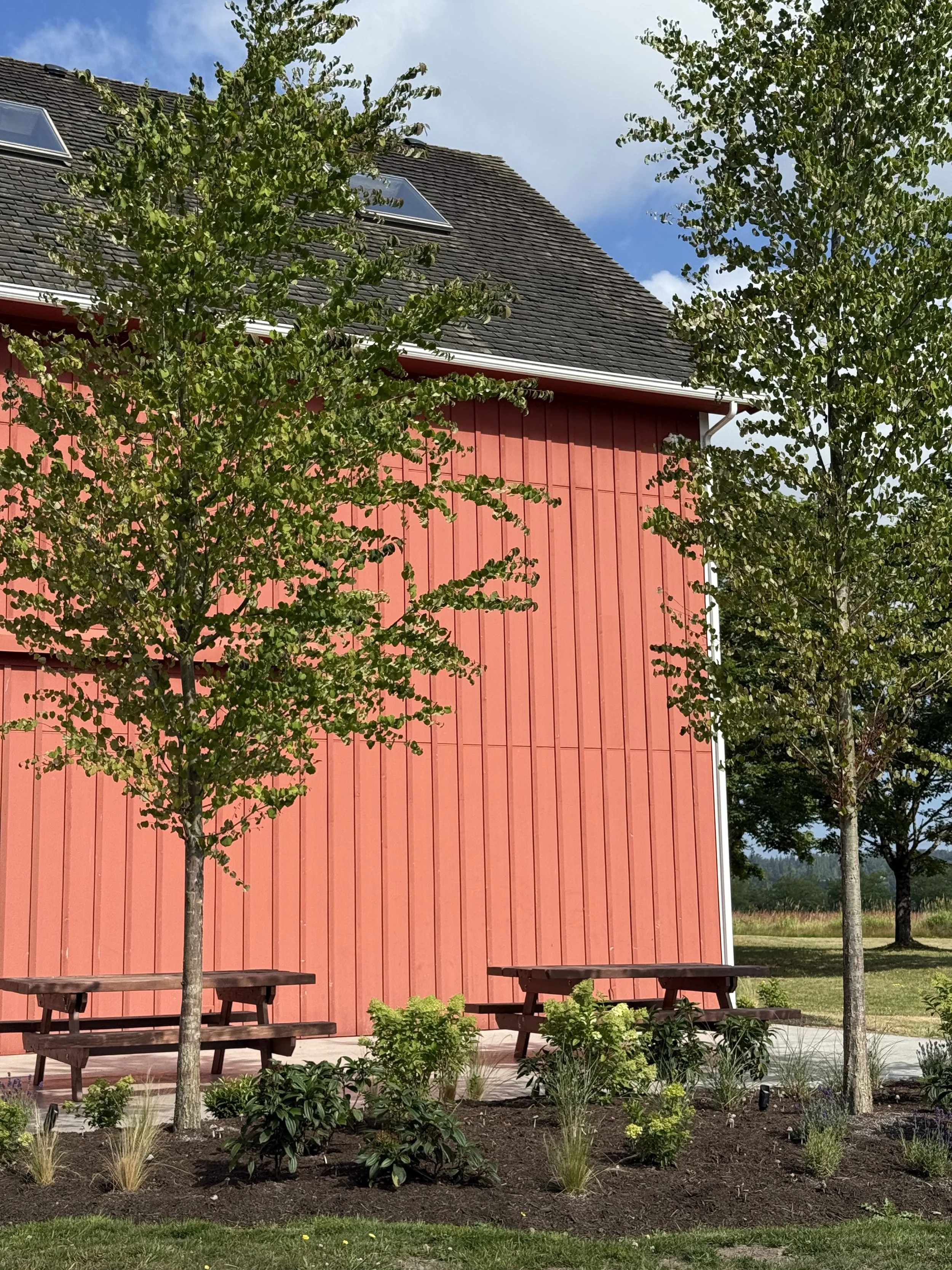Red building with wooden siding, surrounded by trees, with benches and garden plants in the foreground, under a partly cloudy sky.