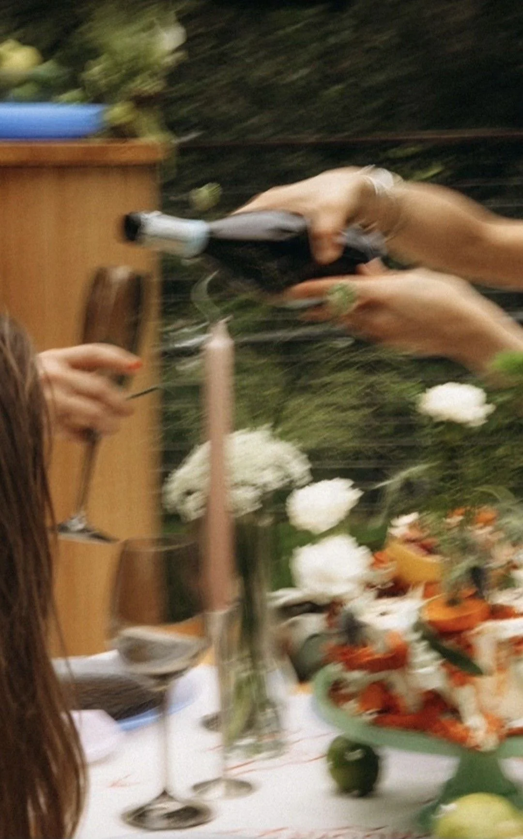 A blurry photo of a person holding a wine bottle and a glass around a table with flowers and food, possibly at a party or celebration.