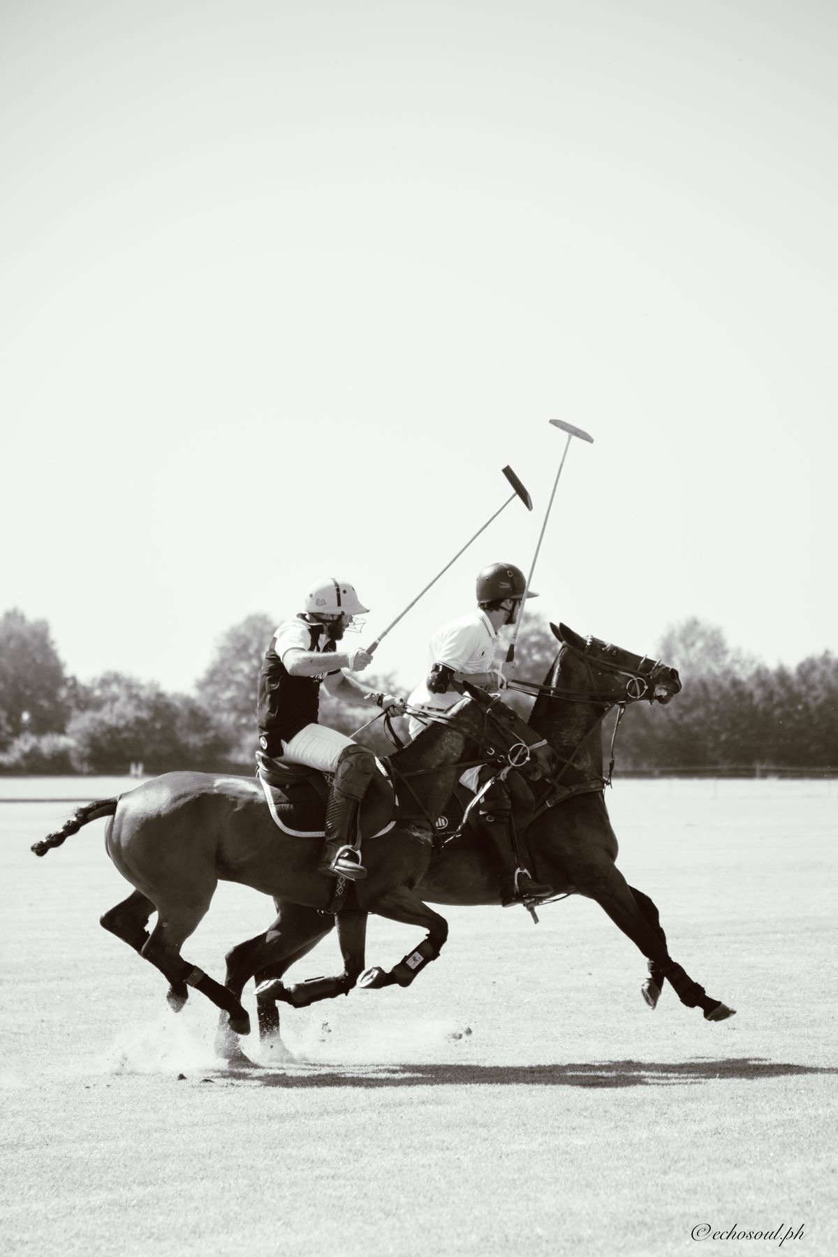 Two polo players on horseback, one in front with a mallet, the other behind, both wearing helmets, in black and white photo.