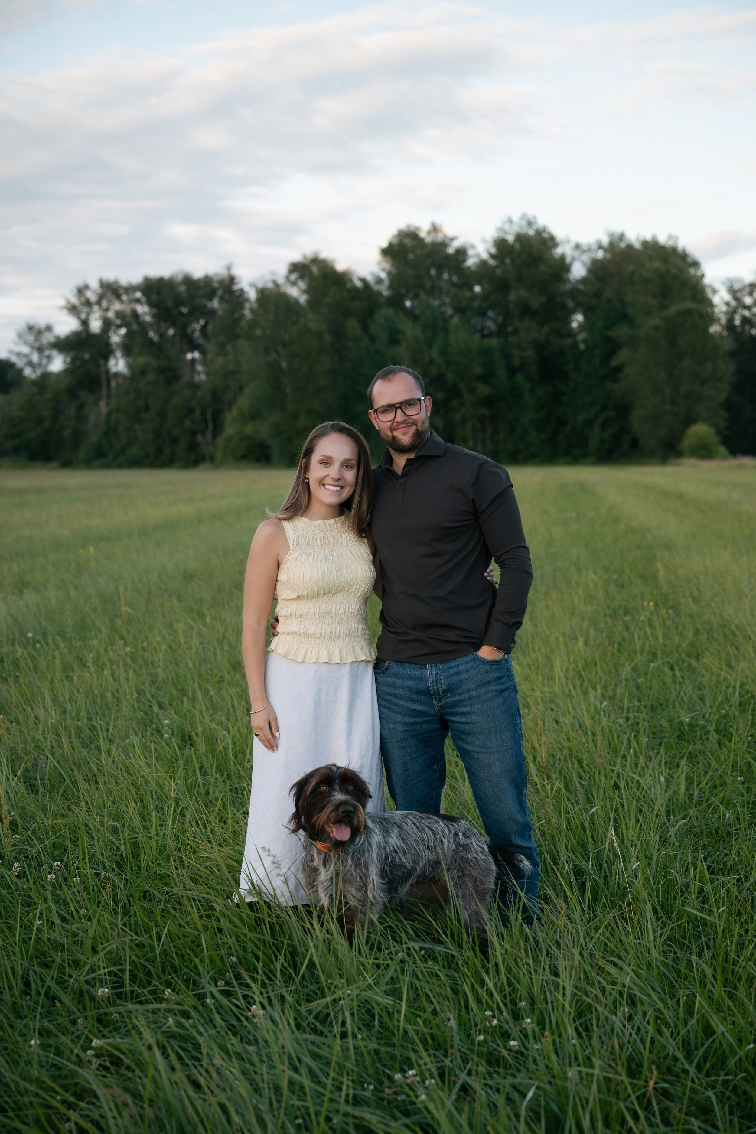 A smiling woman in a cream-colored sleeveless top and white skirt, and a man in a black long-sleeved shirt and blue jeans, standing in a grassy field with trees in the background, with a large dog with black, brown, and gray fur in front of them.