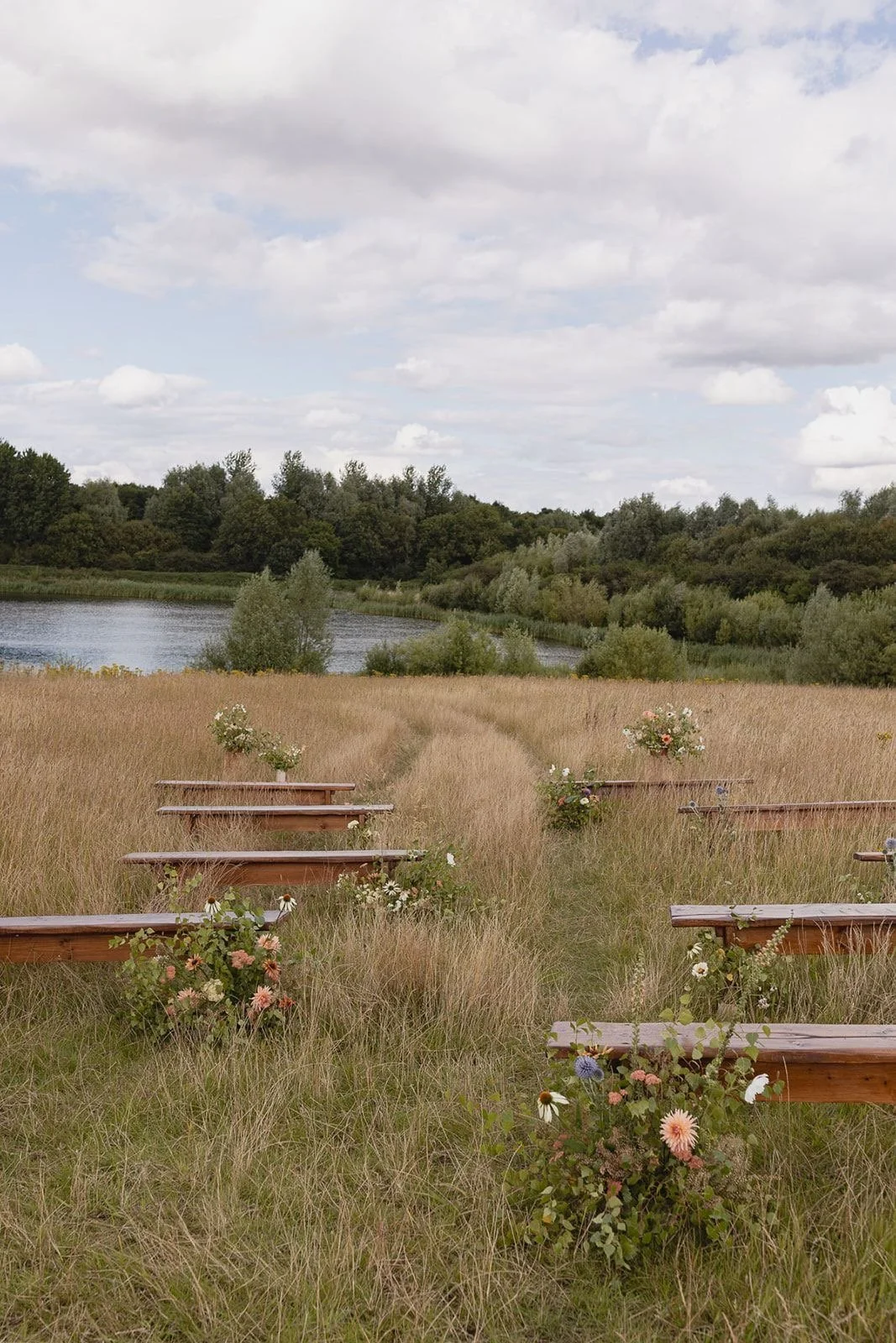 Outdoor wedding or ceremony setup in a grassy field with wooden benches decorated with flowers, overlooking a lake with a forest in the background under a partly cloudy sky.