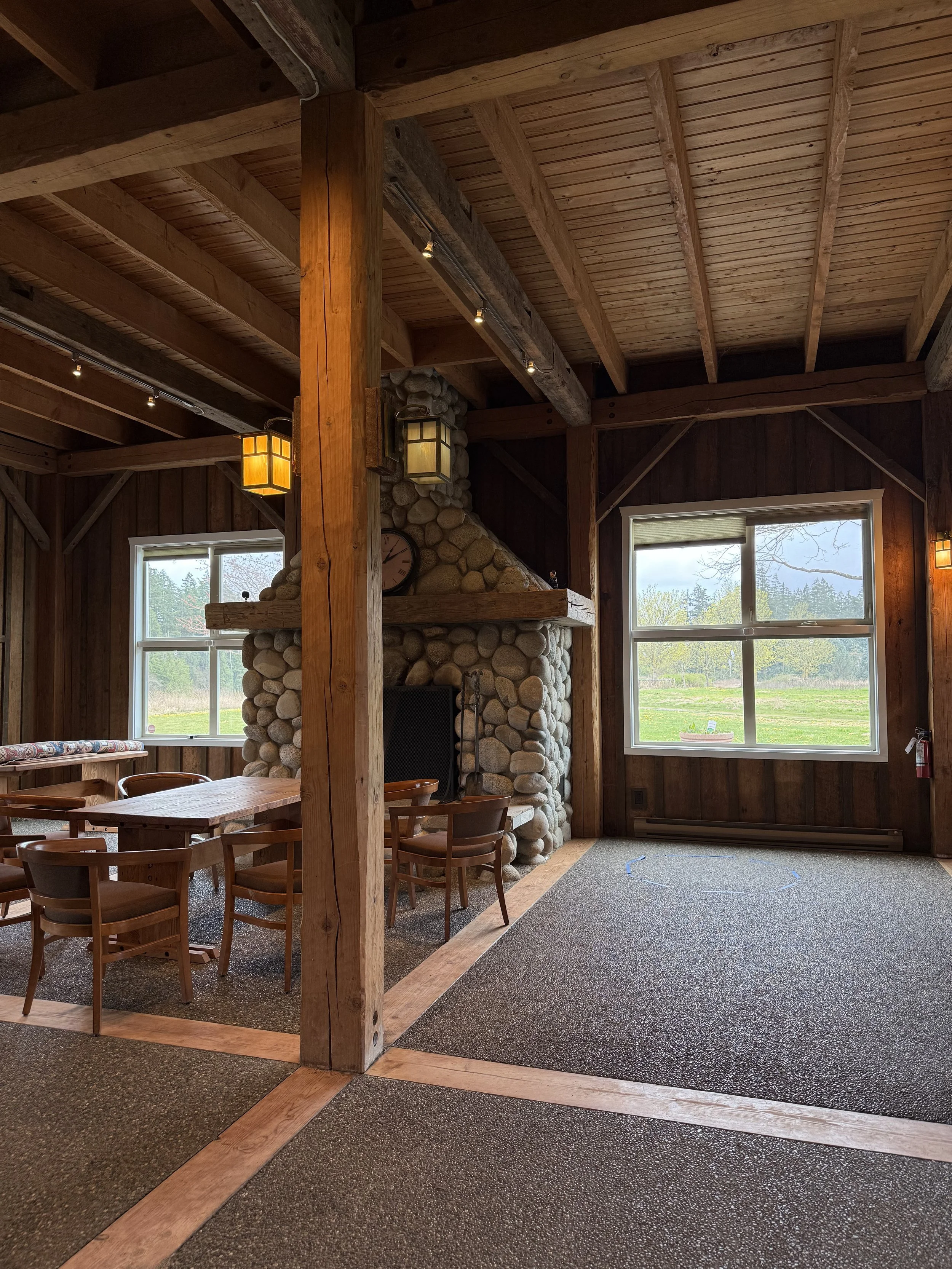 Interior of a rustic wooden lodge with exposed beams and a stone fireplace, large windows showing trees and fields outside, and a dining area with chairs and a table.