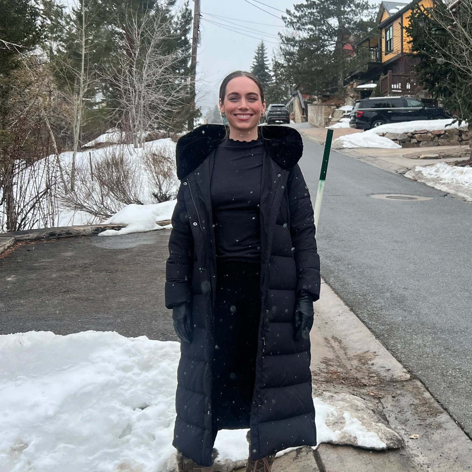 The lead closet and home organizer is standing outdoors on a snowy street, smiling at the camera, dressed in a long black winter coat, gloves, and black pants.