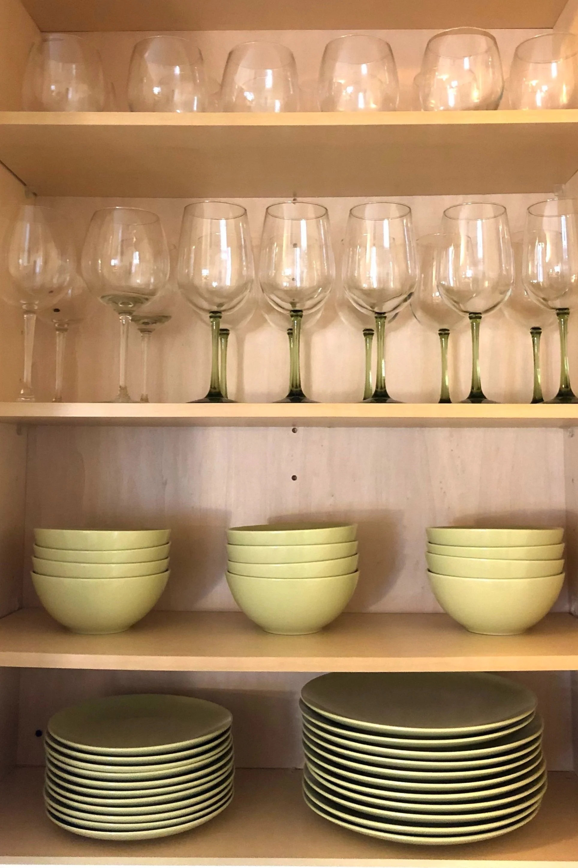 Wooden kitchen cabinet with neatly organized wine glasses on top shelves, stacked green bowls on middle shelf, and stacked green plates on bottom shelf.
