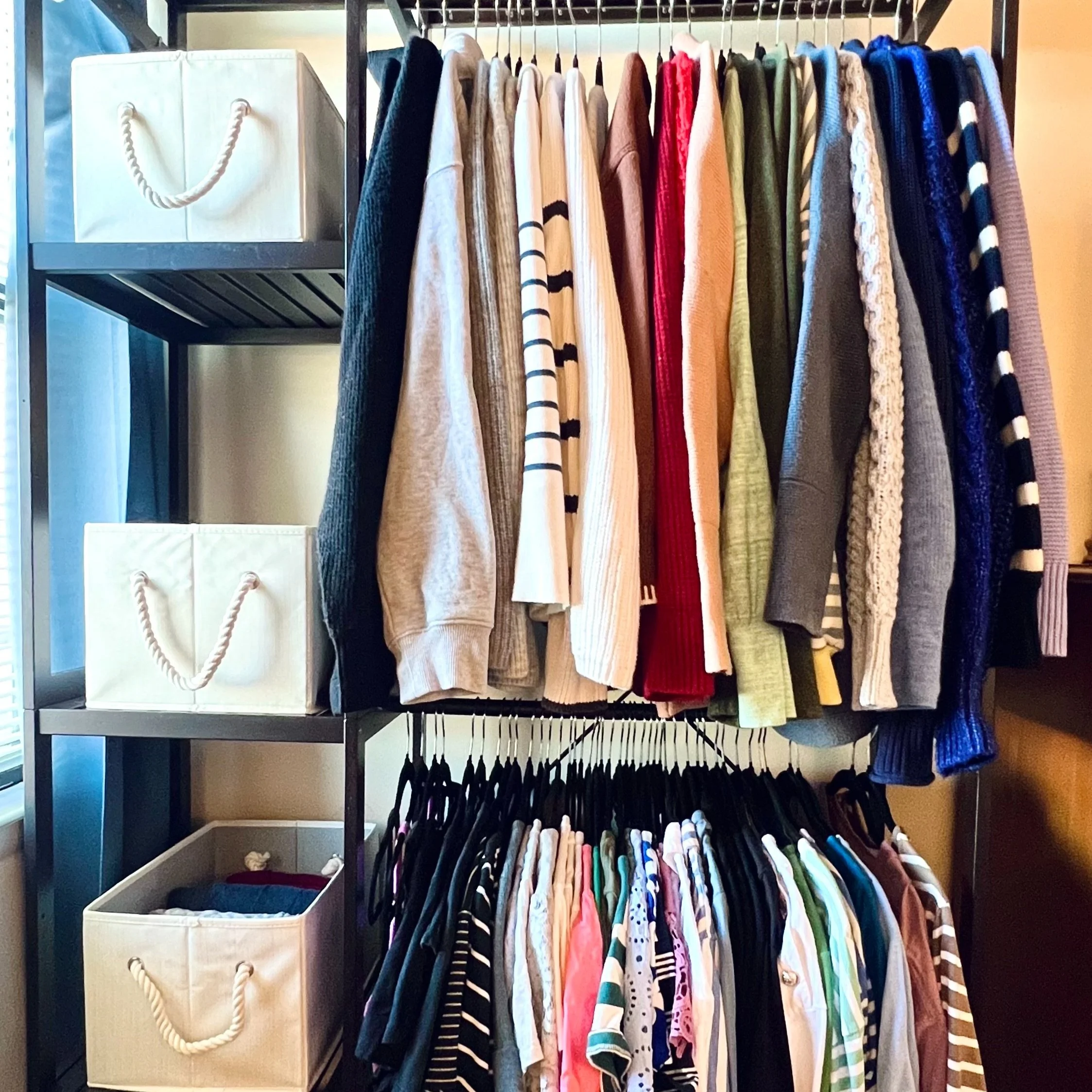 A photo from a closet organizing and stylist session shows a clothing rack with a variety of sweaters and shirts on hangers, with two white storage boxes on a black metal shelf beside it.