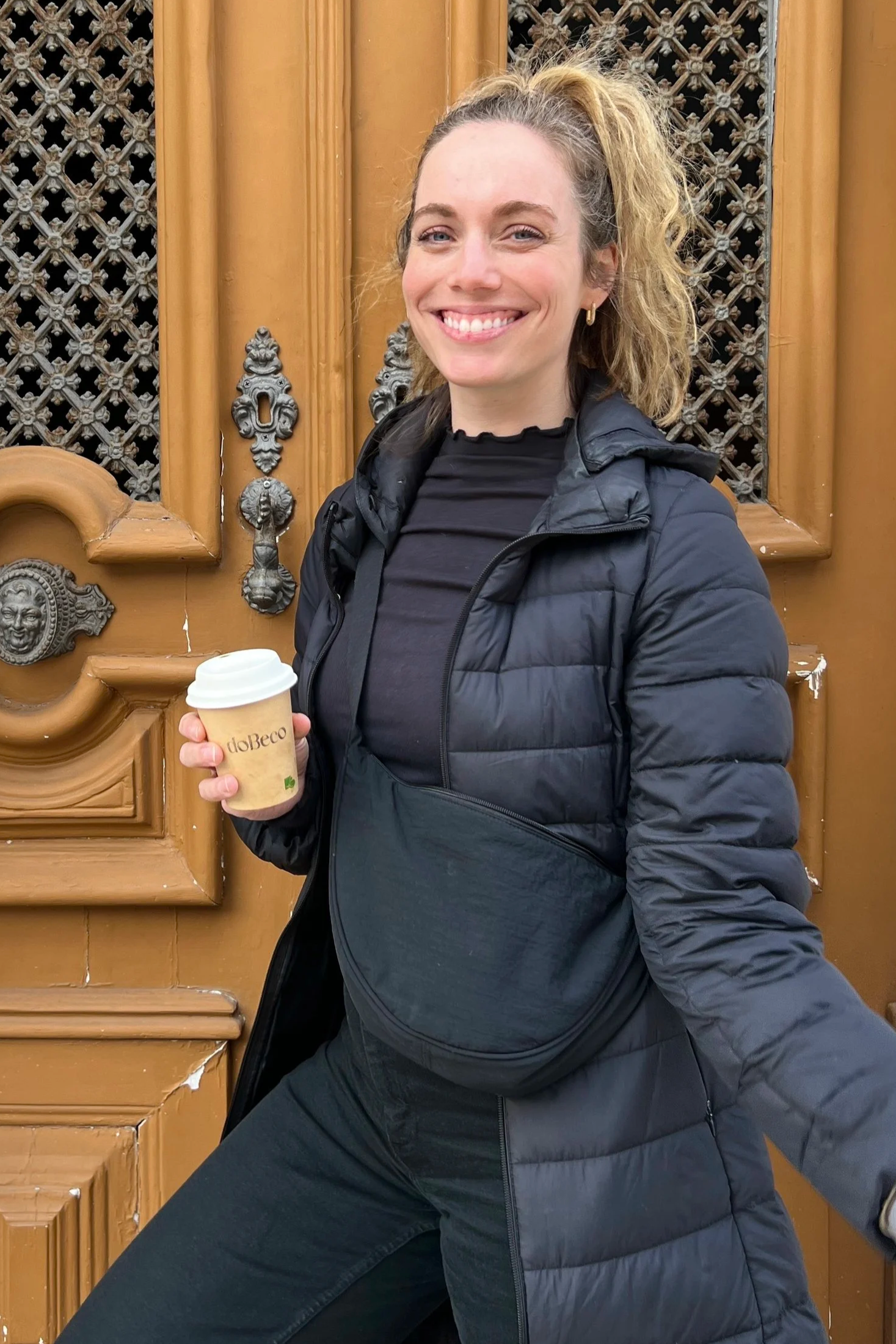 The lead closet and home organizer is smiling and holding a paper coffee cup, standing in front of an ornate wooden door with decorative metalwork.