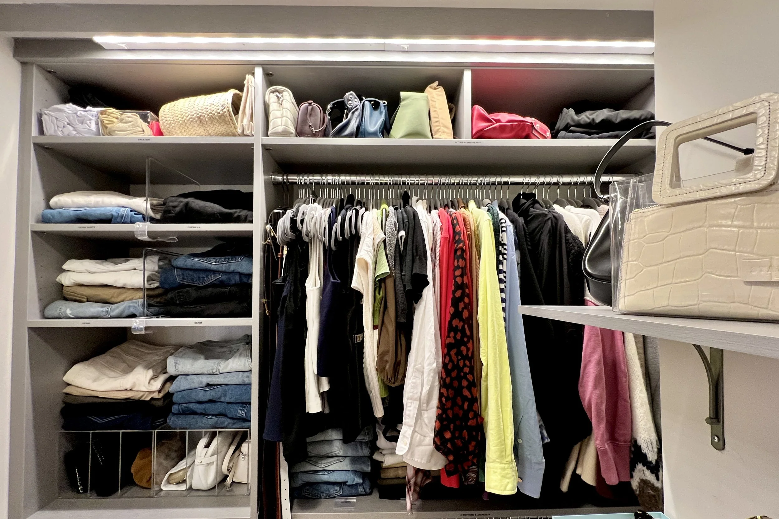 A photo from a closet organizing and stylist session shows a closet with shelves containing folded clothes, handbags, and purses. A hanging rod with various shirts, blouses, and jackets. On the right, a beige crocodile-patterned handbag on a shelf.