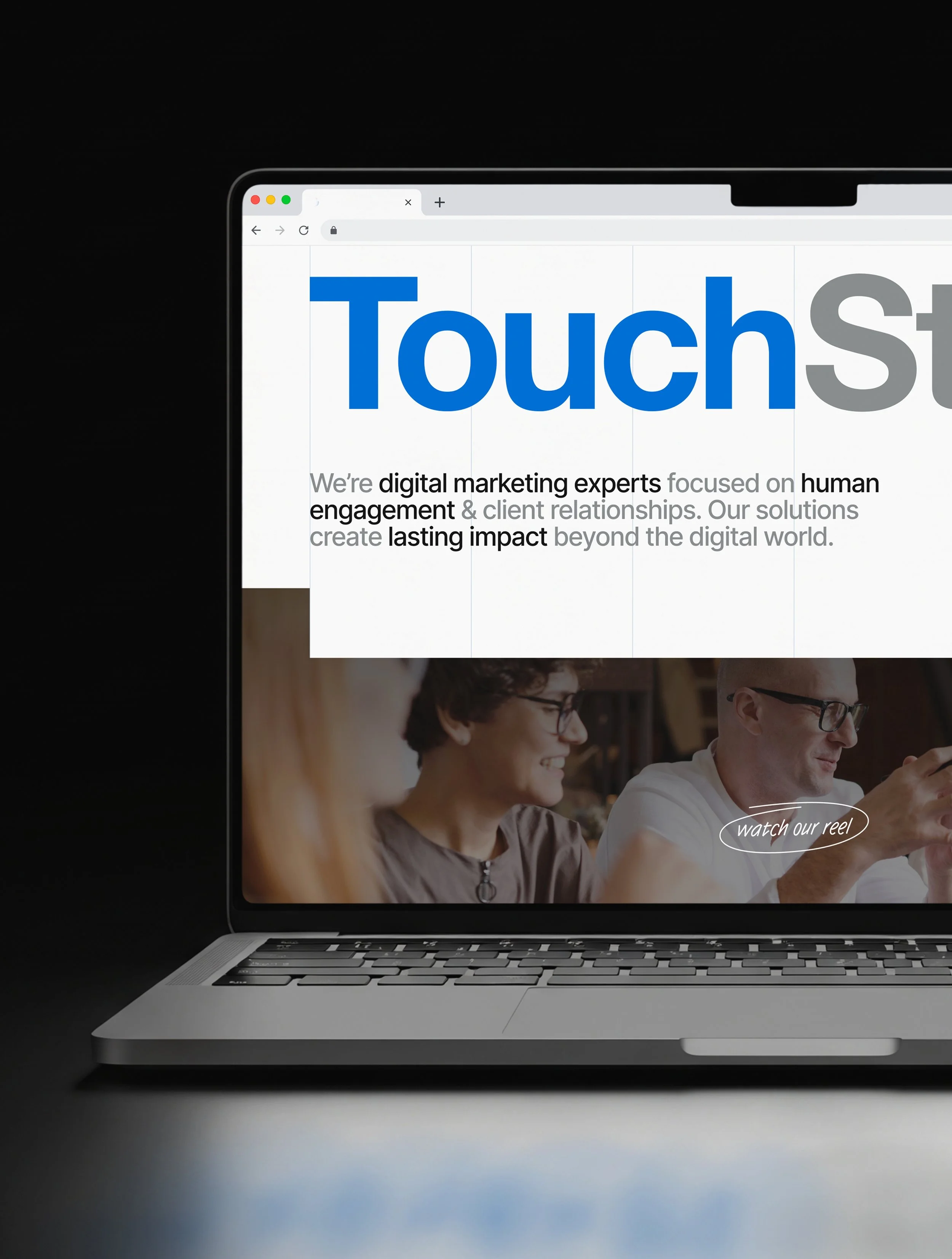 Close-up of a silver MacBook laptop on a white surface against a black background, displaying a website with the word 'Touch' in large blue letters and a photo of two people smiling.