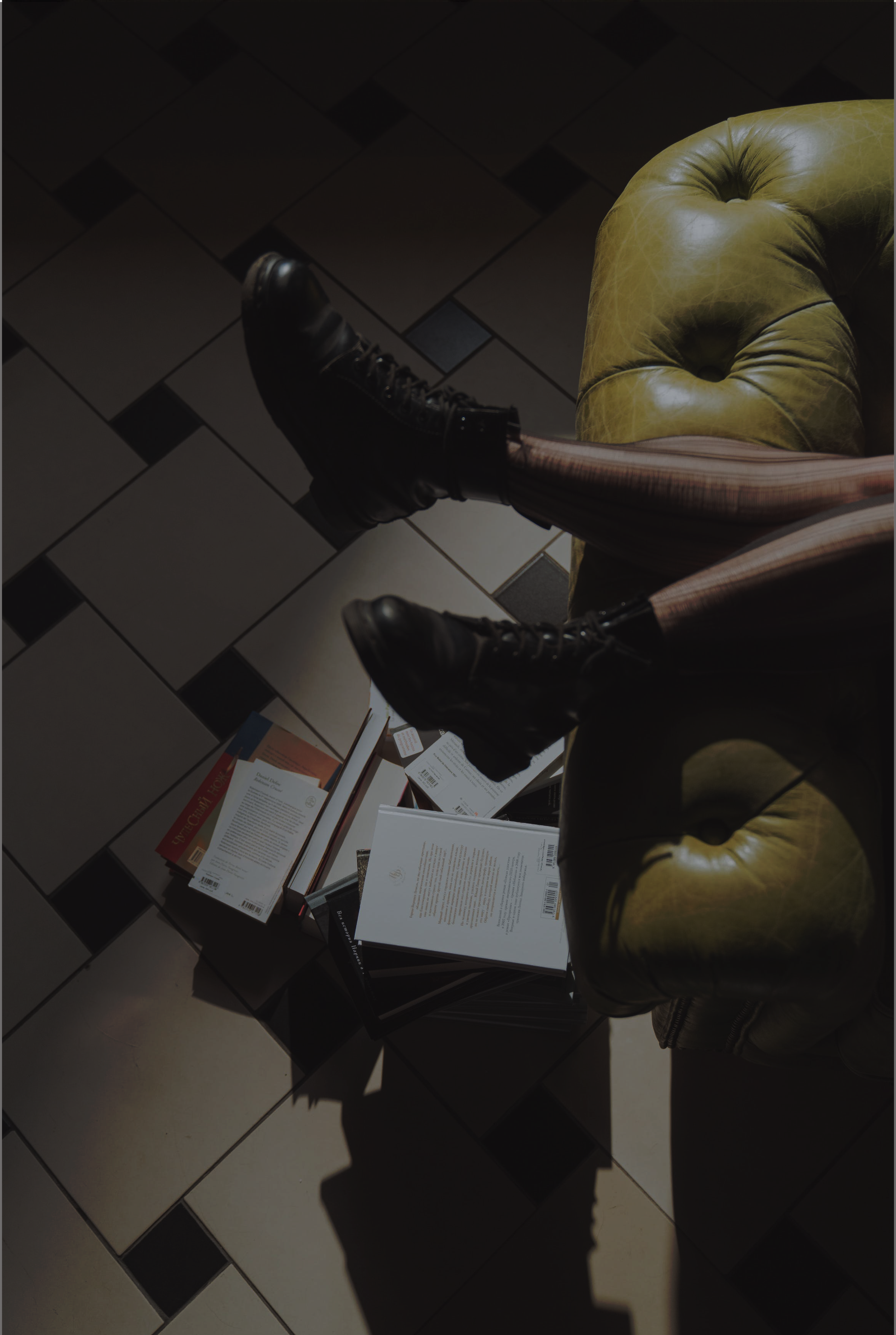 View of a tiled floor with a green leather armchair, a person wearing black boots and striped pants sitting on it, and a table with books and papers.
