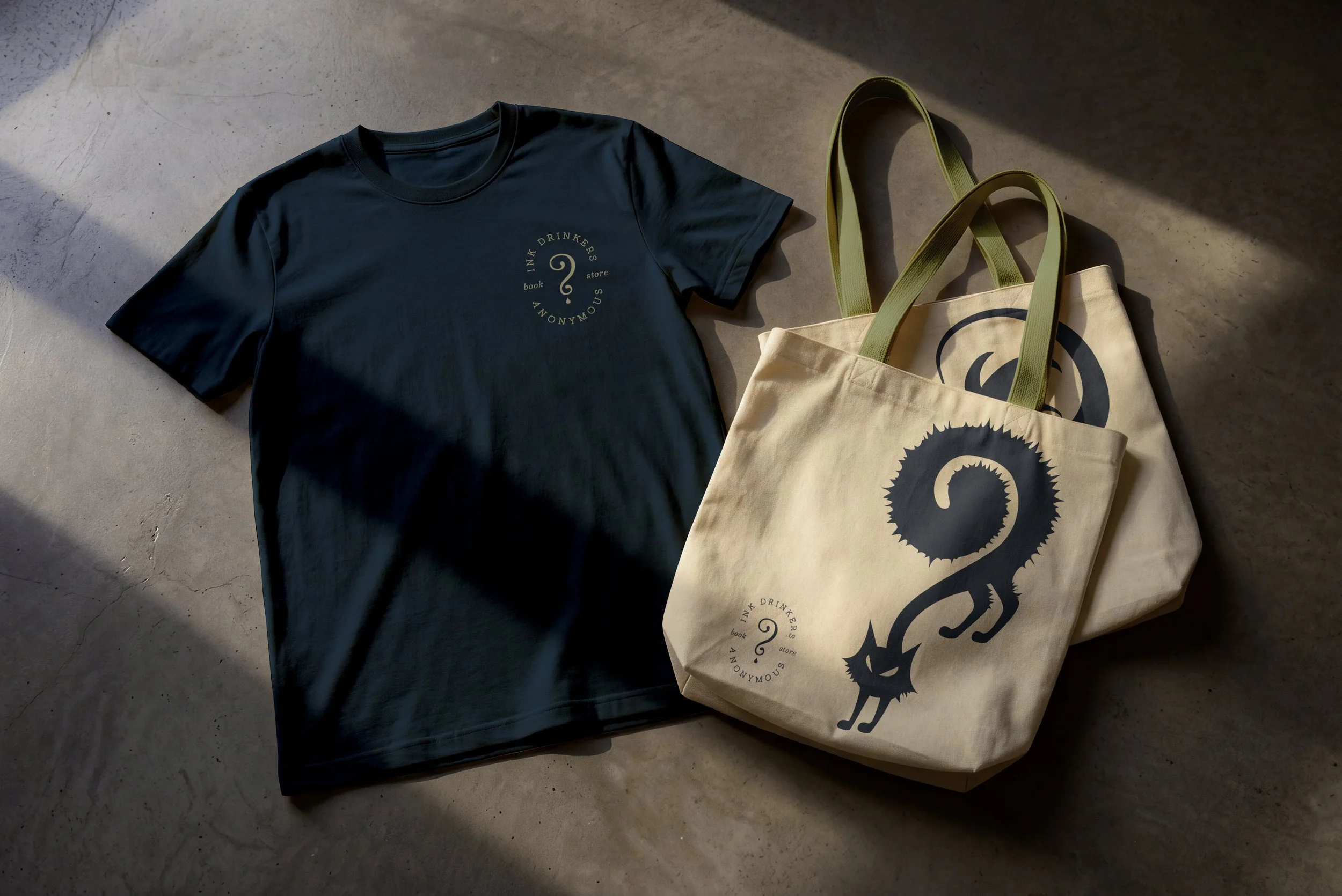A black T-shirt with a circular logo in the center reading 'INK DRINKERS BOOK STORE ANONYMOUS' with a question mark in the middle, and a cream-colored tote bag with a black dragon design and additional similar circular logo, both placed on the ground with shadows cast on the surface.