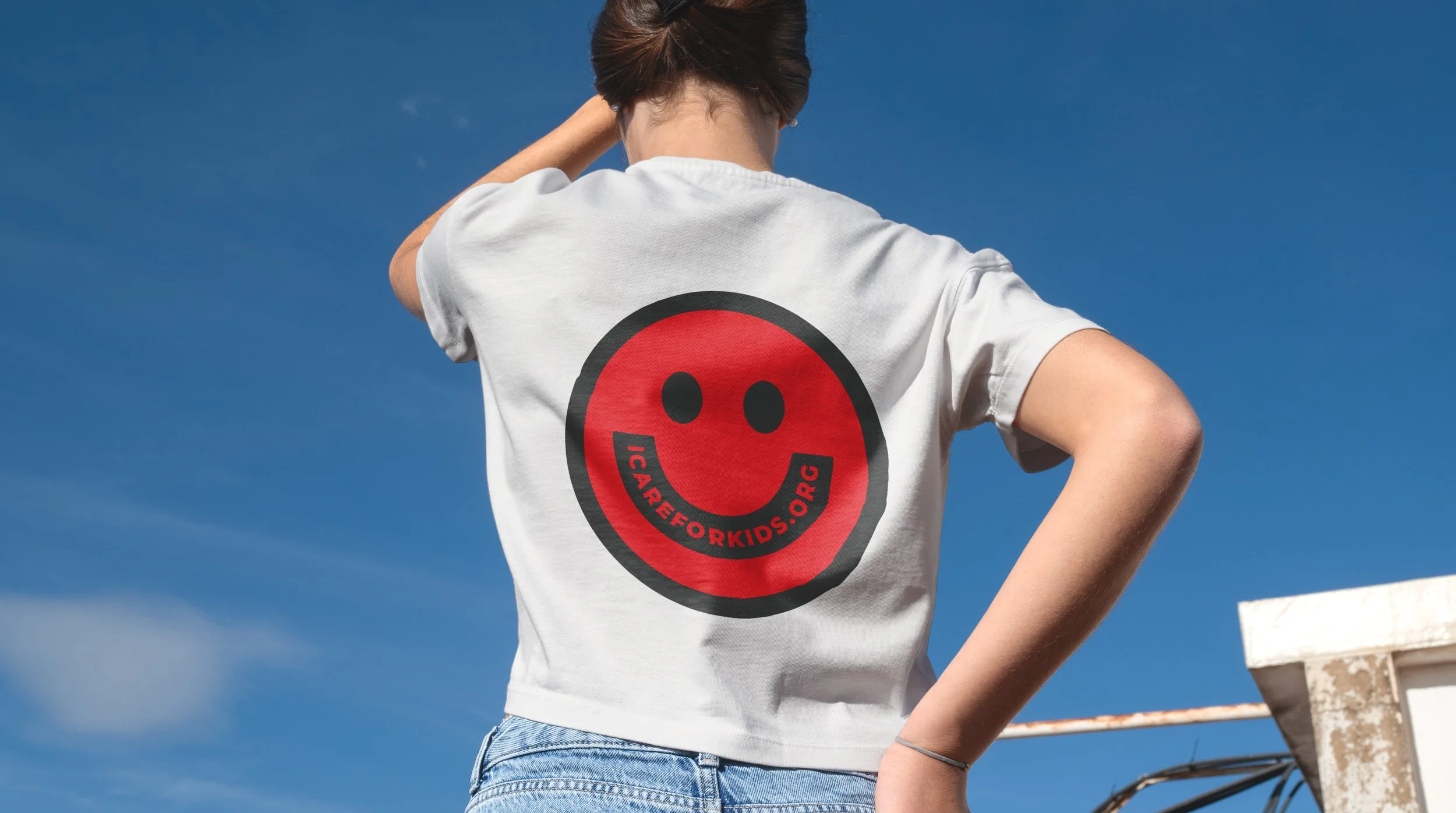 Person with short brown hair wearing a white T-shirt with a red smiley face logo and the words 'ICAREFORKIDS.ORG' on the back, standing outdoors against a blue sky.