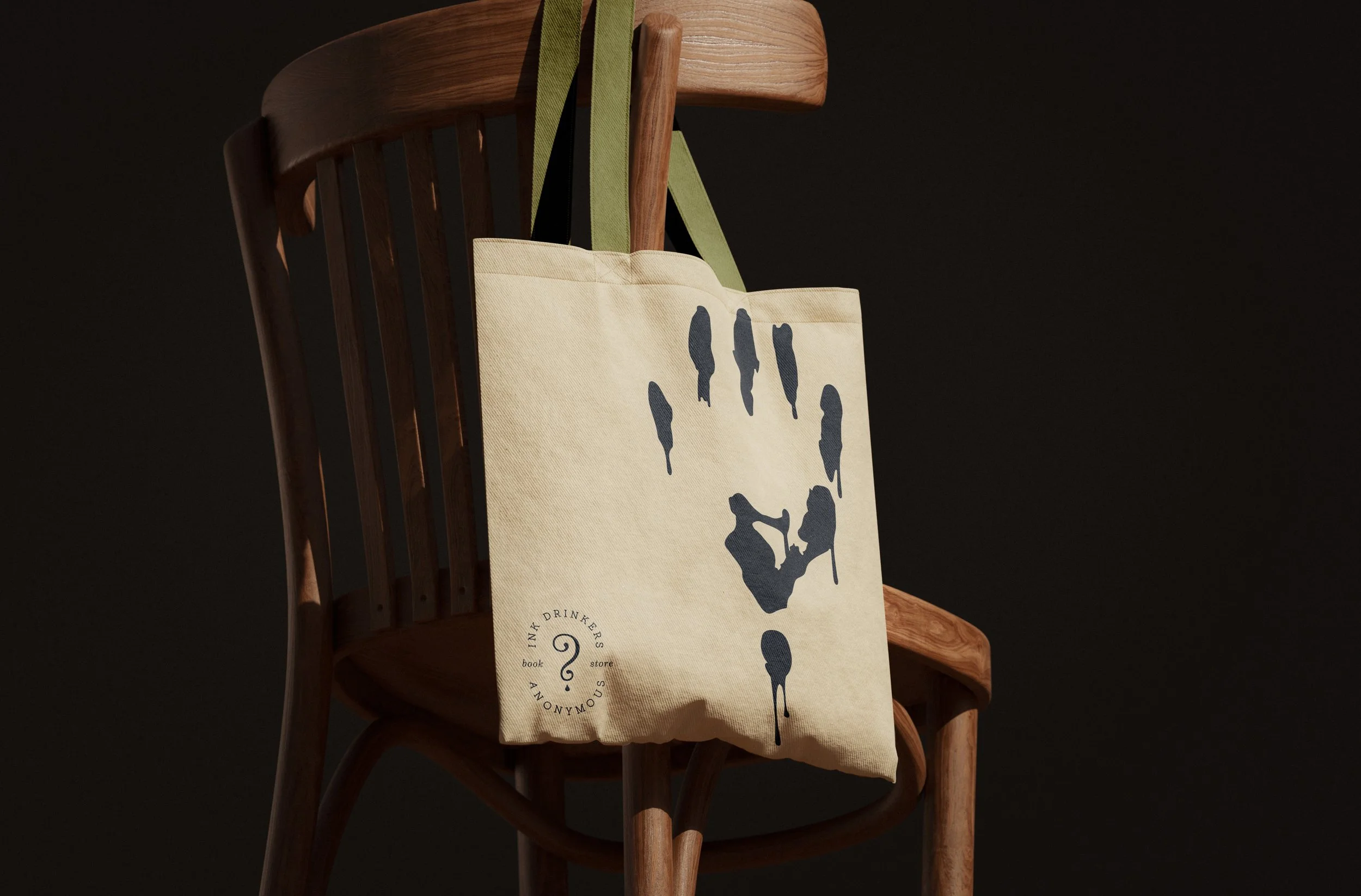 A wooden chair with a beige tote bag hanging on it, featuring black and green straps. The bag has a dark graphic design and text that reads 'SOU' and 'IN. MAK DRINKERS' with a circular logo.