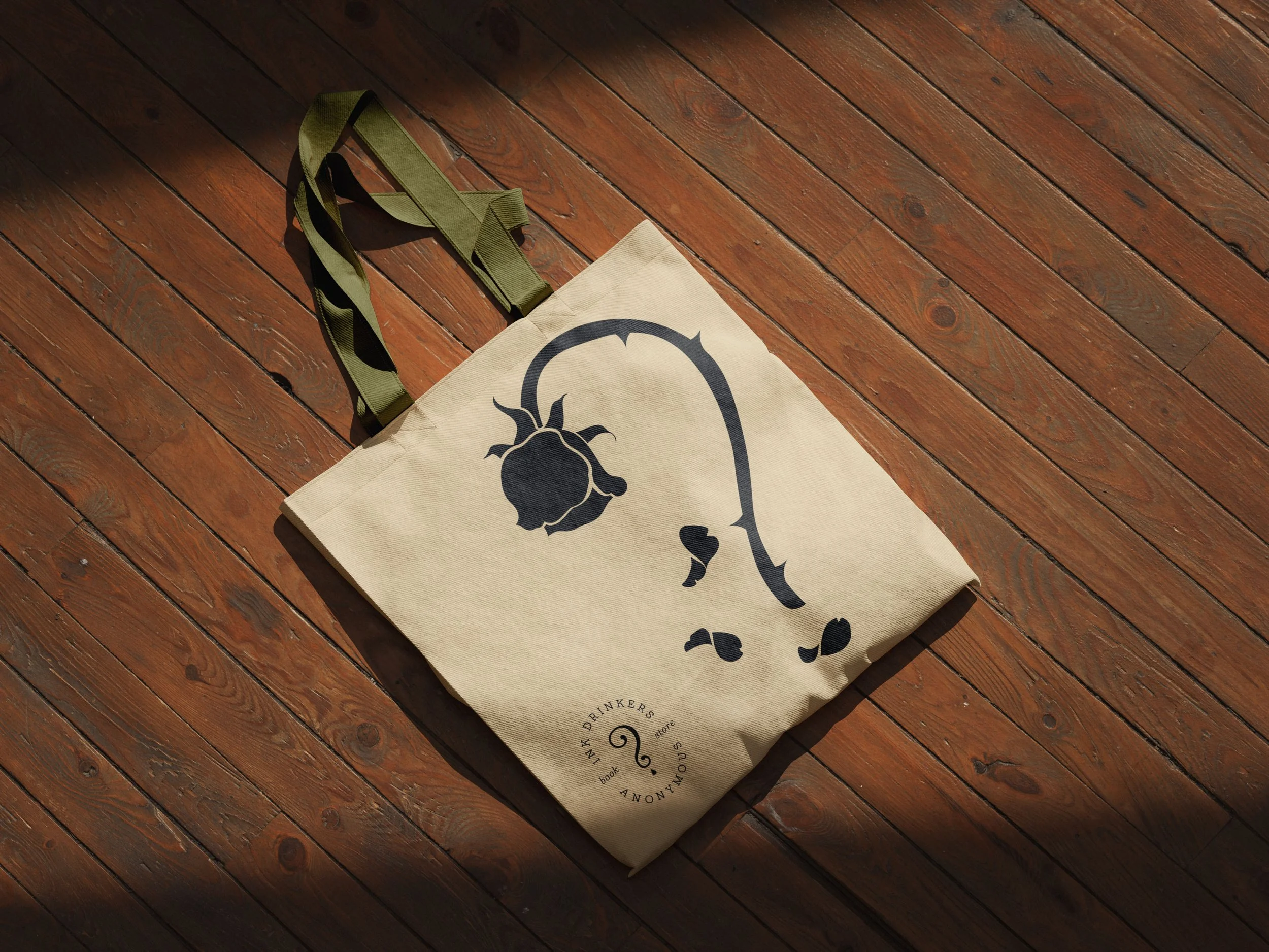 A beige tote bag with a black beetle graphic lying on a wooden floor, with a green strap attached.