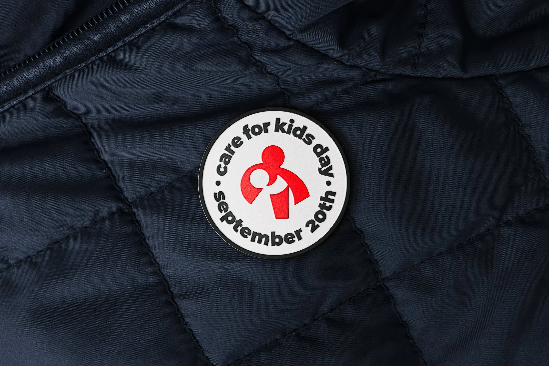 Close-up photo of a black quilted fabric jacket with a circular badge that reads 'care for kids and' with a red symbol in the center.
