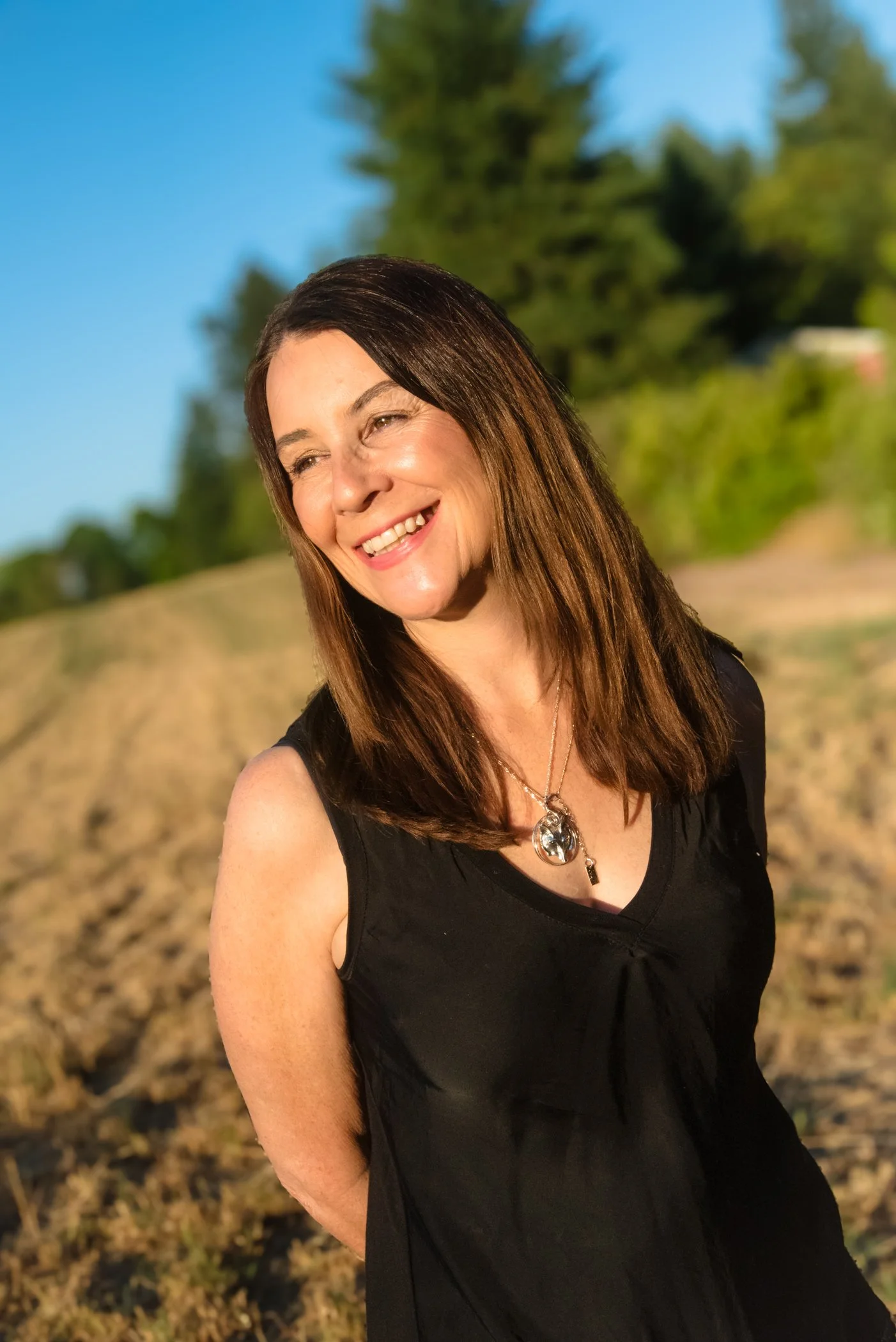 Alison Potts smiling outdoors in a black sleeveless top with long brown hair and a necklace, standing in front of a field and trees under a clear blue sky.