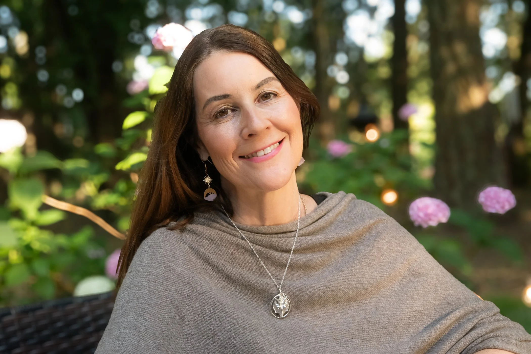 A woman with long brown hair smiling outdoors, wearing earrings, a necklace, and a gray top, with blurred greenery and pink hydrangea flowers in the background.