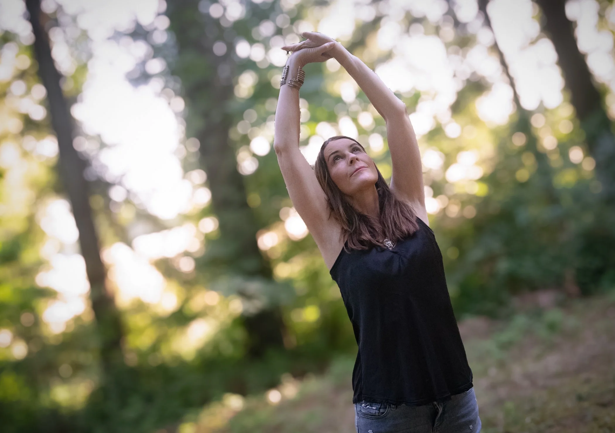 Alison Potts, wearing a black sleeveless top and jeans, standing outdoors in a wooded area with sunlight filtering through trees, reaching with both hands above her head and looking upward.