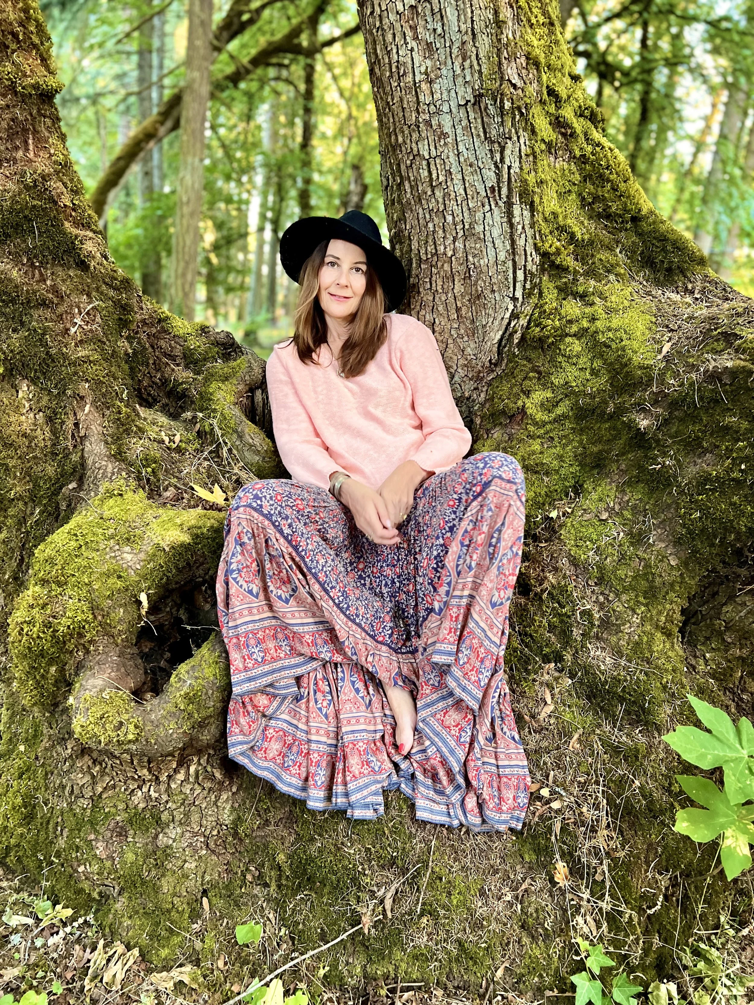 Alison Potts sitting against a large moss-covered tree in a forest, wearing a pink sweater, patterned skirt, and a black wide-brimmed hat.
