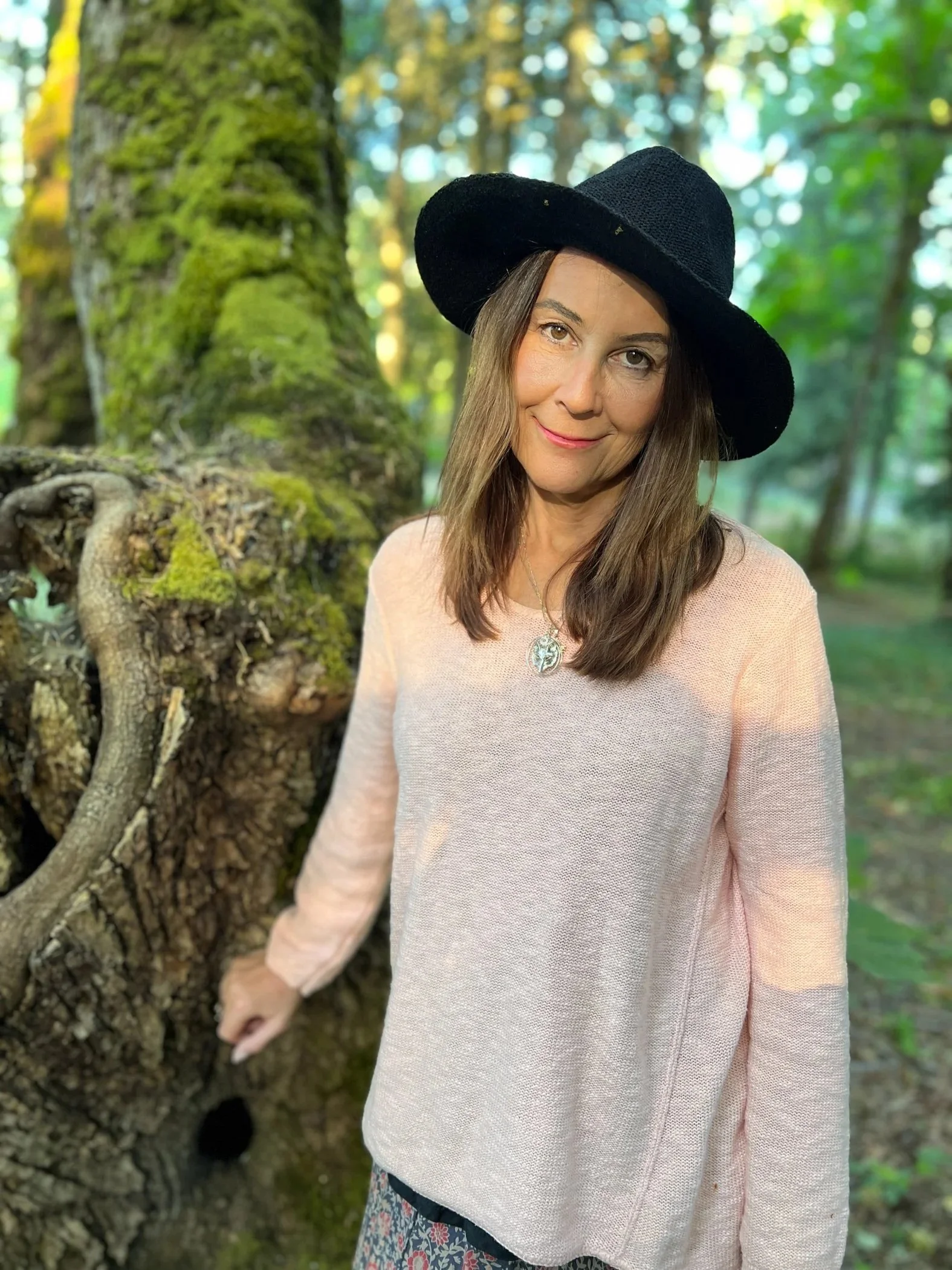 Alison Potts wearing a black wide-brimmed hat and a light pink sweater, standing outdoors in a forested area with green foliage and a large tree trunk covered in moss.