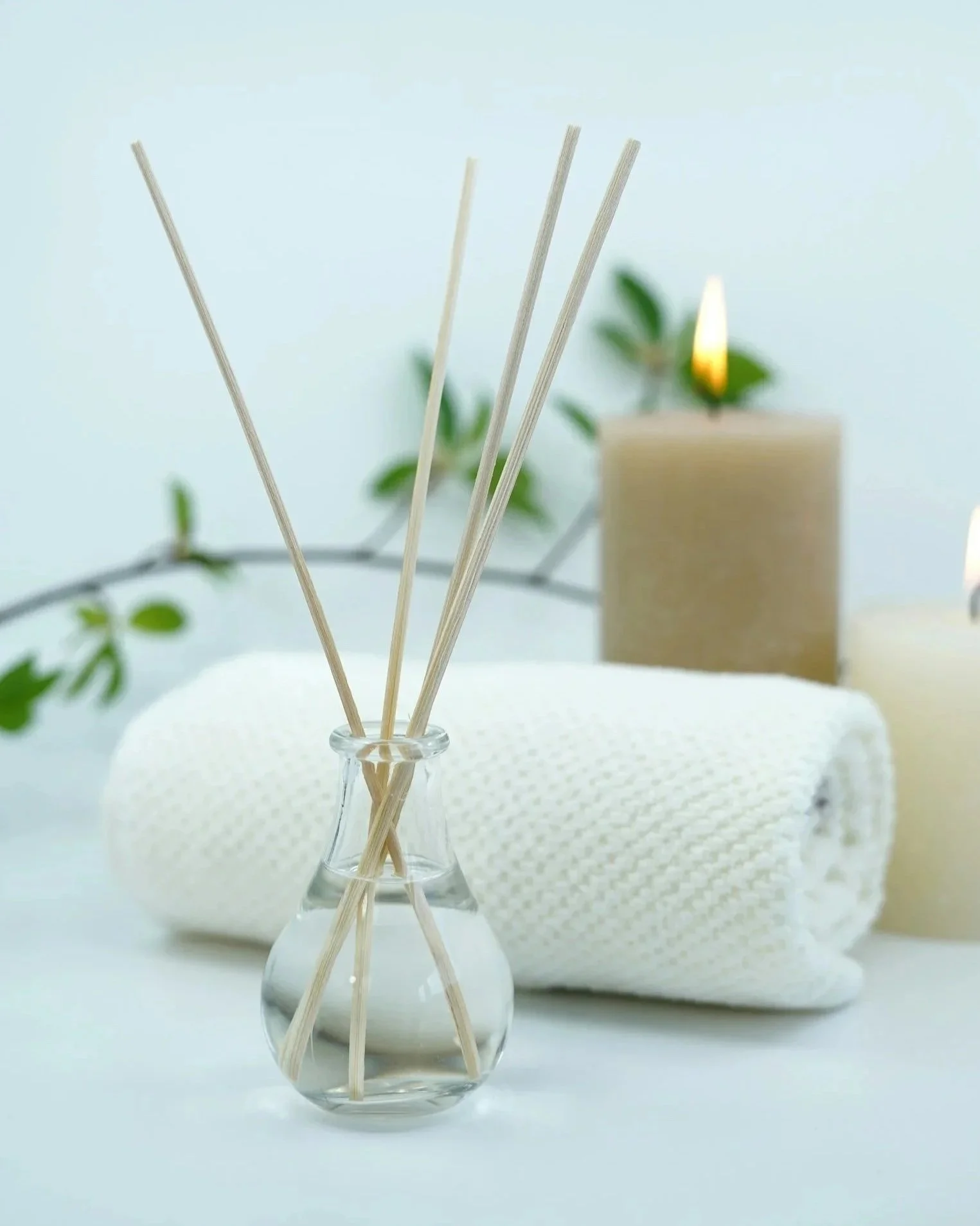 Reed diffuser in a small glass vase on a white surface, with candles, a rolled white towel, and greenery in the background.