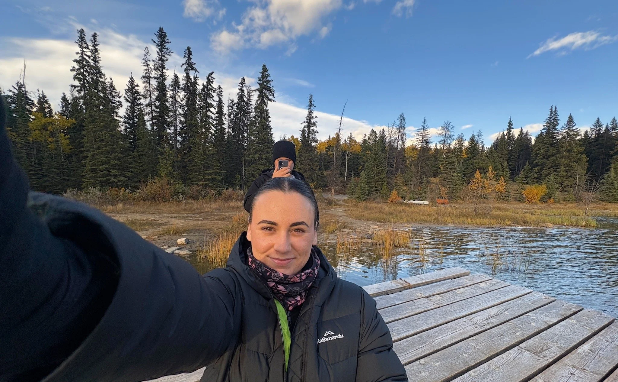 Two women taking a selfie outdoors by a lake in a forested area during autumn. One woman in the foreground is smiling, wearing a black jacket and a patterned neck gaiter. The other woman in the background is using her phone, wearing a black beanie and jacket. There is a dock extending into the water and trees with fall foliage under a blue sky with some clouds.