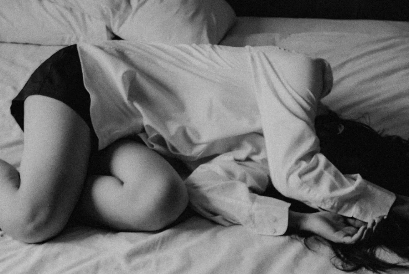 A person lying on a bed curled up with their arms around their knees, wearing a light-colored shirt and dark shorts, with their head turned to the side and long hair spread out on the pillow.