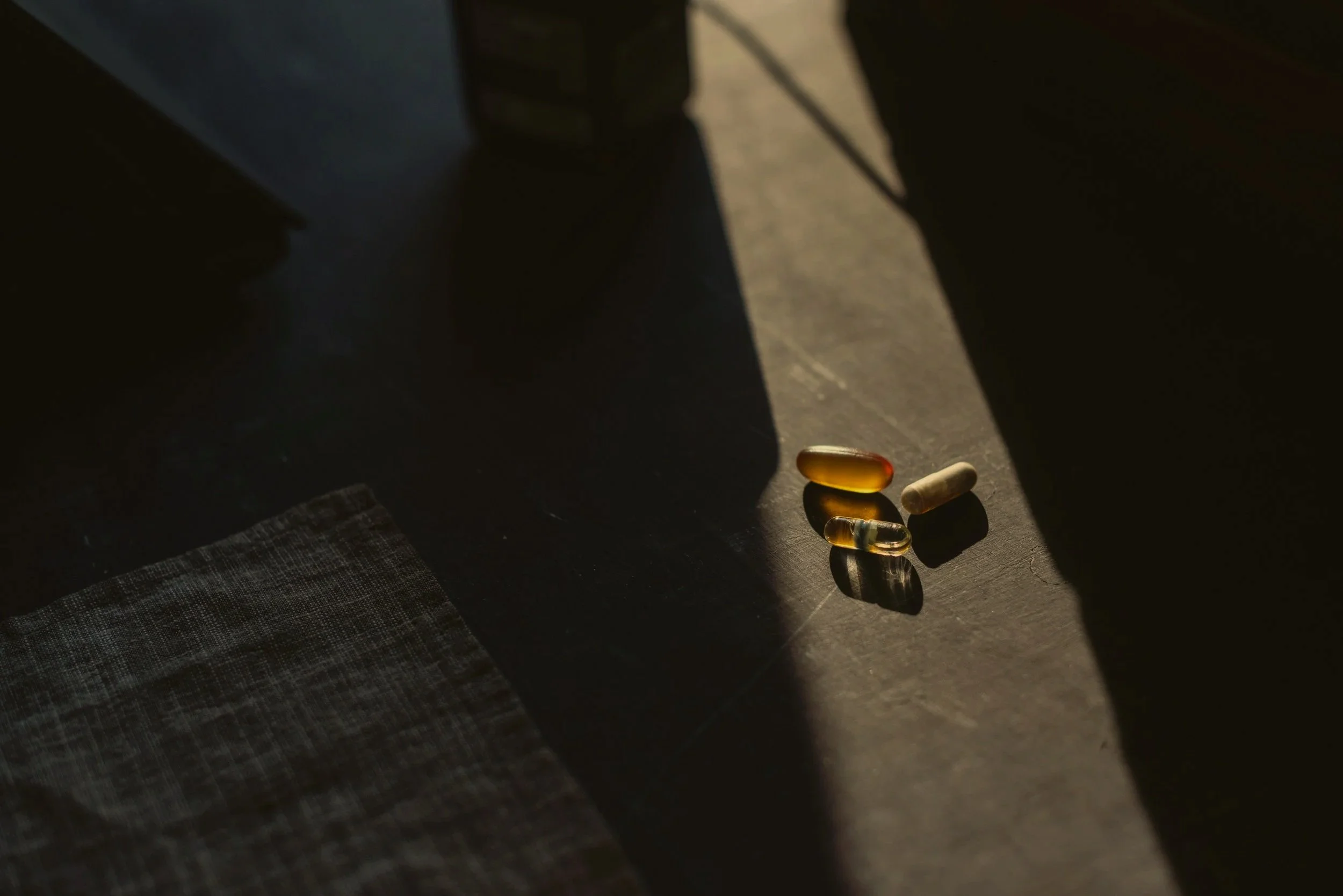Four pills and capsules on a dark surface in sunlight shadows, with part of a cloth at the bottom left.