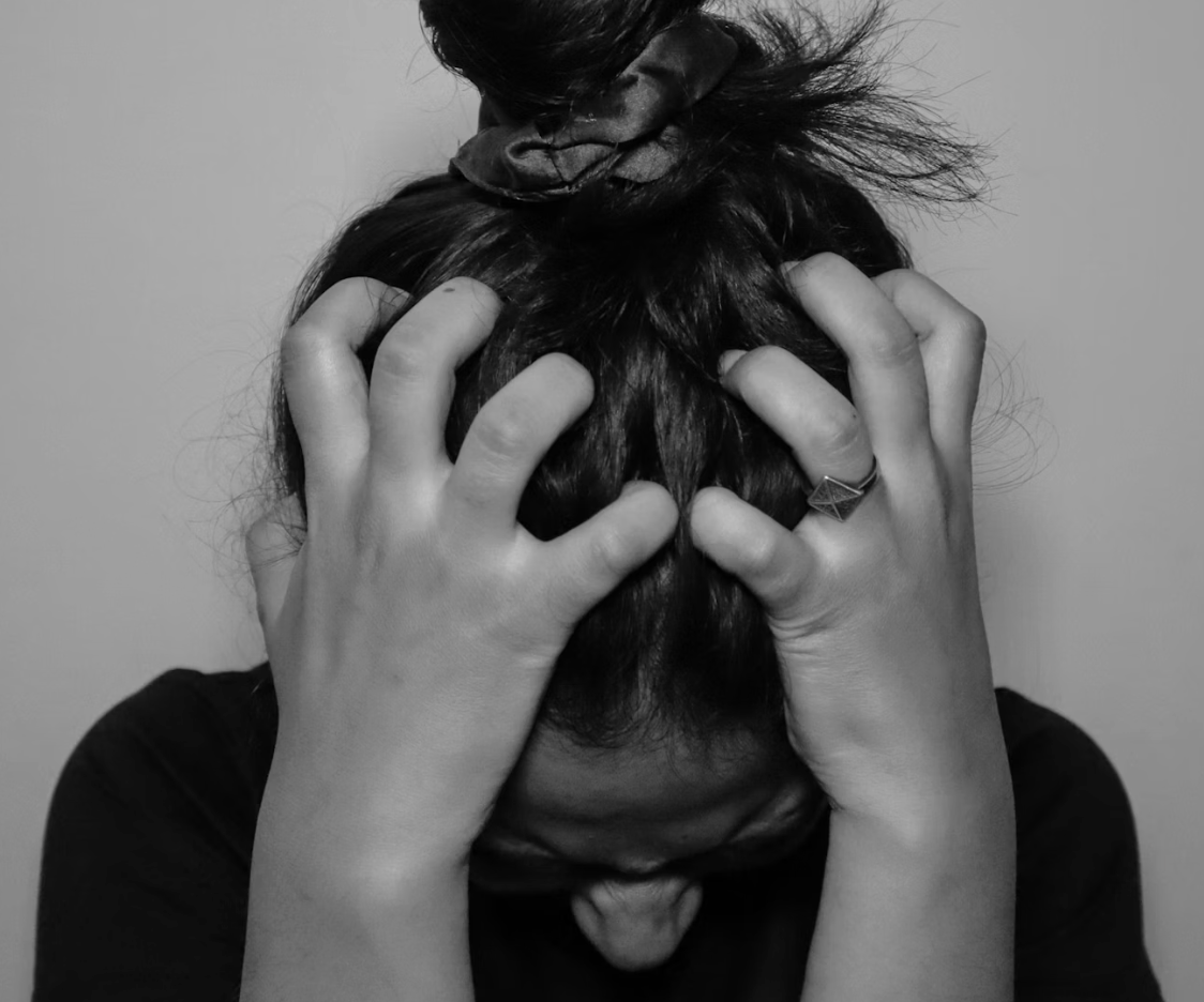 Person with dark hair holding their head with both hands, hair tied up with a scrunchie, in a black and white photograph. In visible pain or anguish.