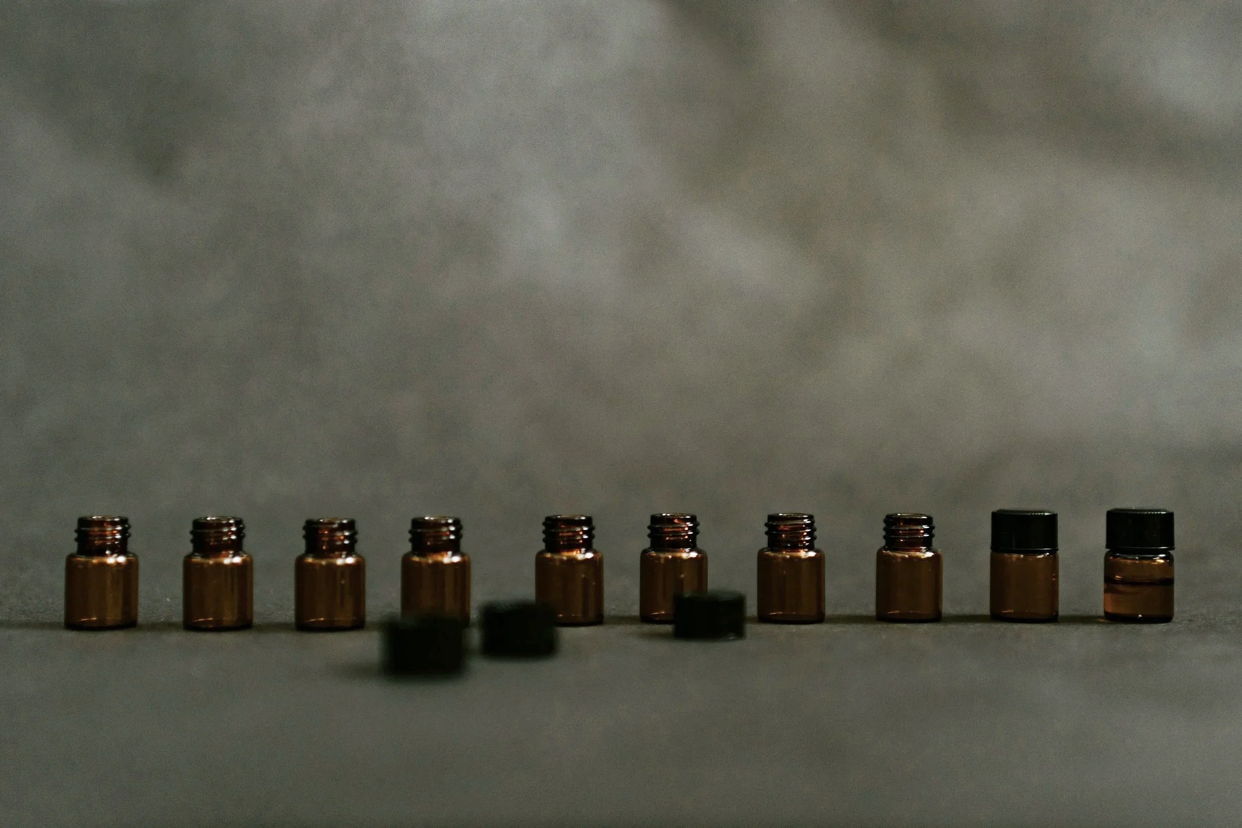 A row of small amber glass bottles with black caps, some of which are capped and some are open, placed on a flat surface against a gray background.