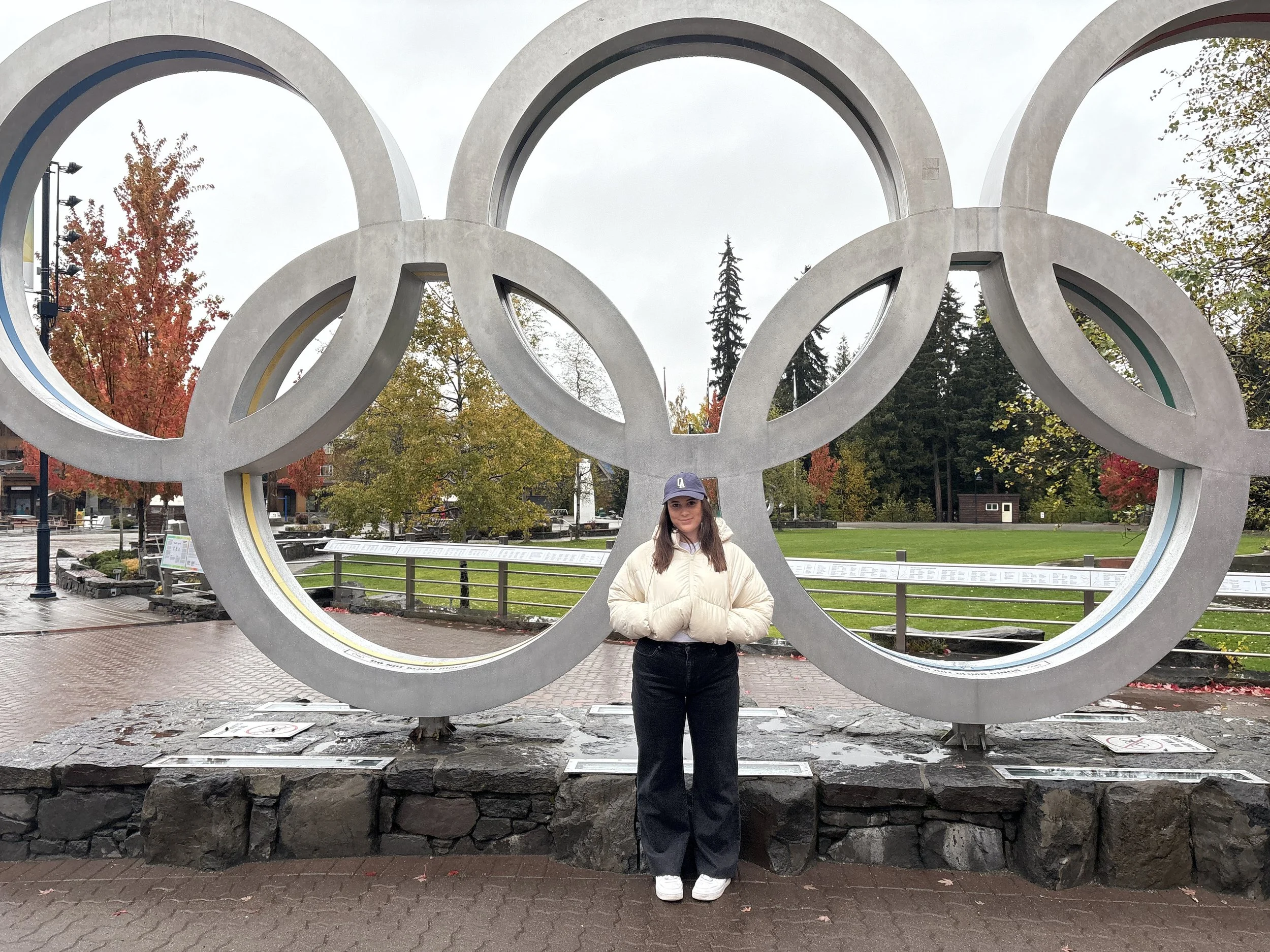 A woman standing in front of a large Olympic rings sculpture outdoors on a cloudy day, with trees displaying autumn colors in the background.
