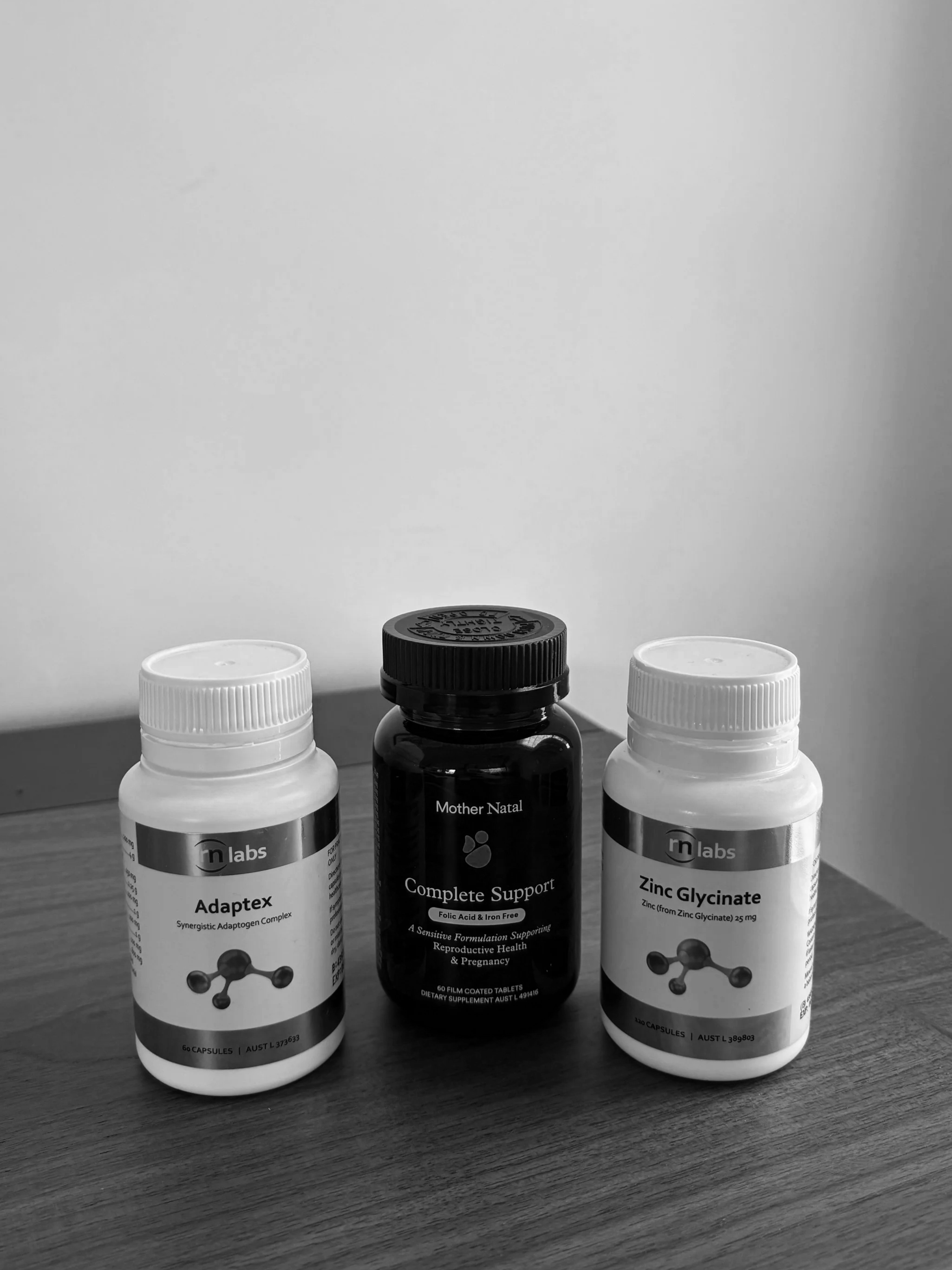 Three bottles on a wooden surface, two white supplement bottles labeled 'Adaptex' and 'Zinc Glycinate' and a black bottle labeled 'Complete Support' from Mother Natal.