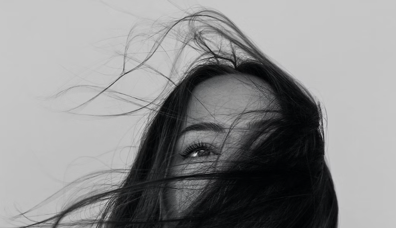 Close-up of a woman's face partially covered by her flowing dark hair, with visible eye and eyebrow, against a plain background.