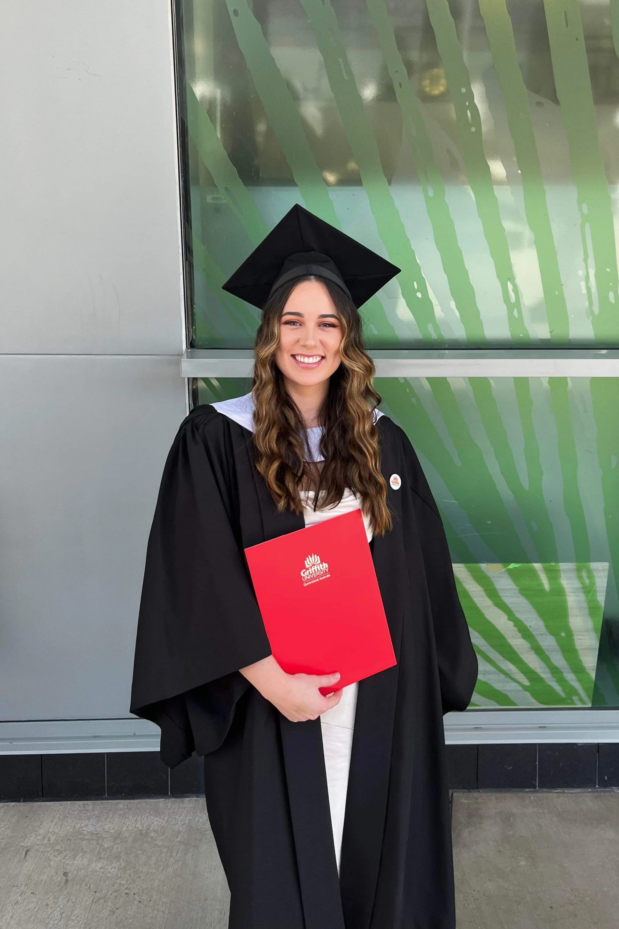A young woman with wavy brown hair smiling in a graduation cap and gown, holding a red diploma folder with a college logo, standing outside in front of a green and gray geometric background.