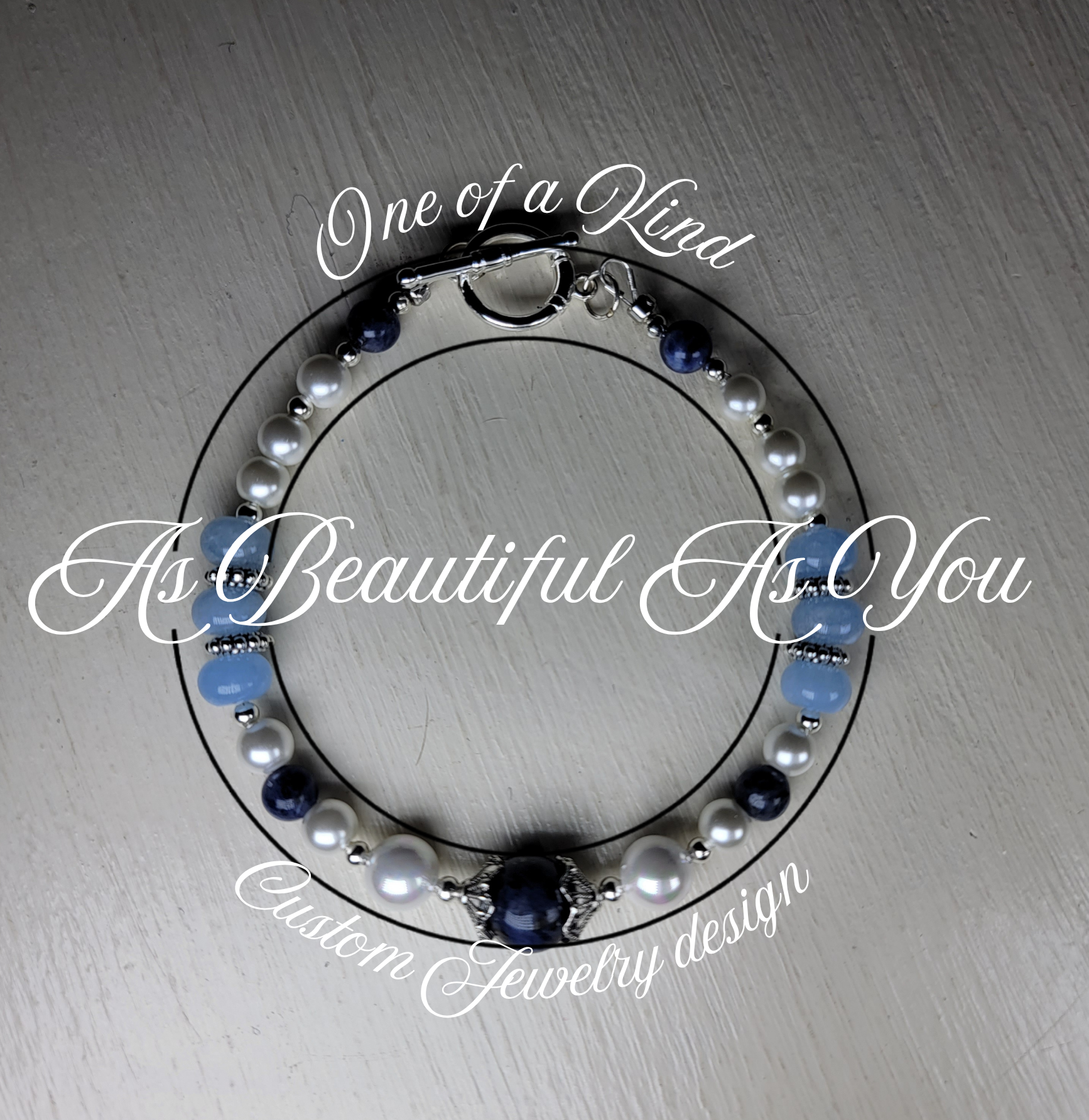 Close-up of a beaded necklace with blue and white pearls, black beads, and silver accents on a light-colored surface, with overlaid white scripted text that reads 'One of a Kind, As Beautiful As You'.