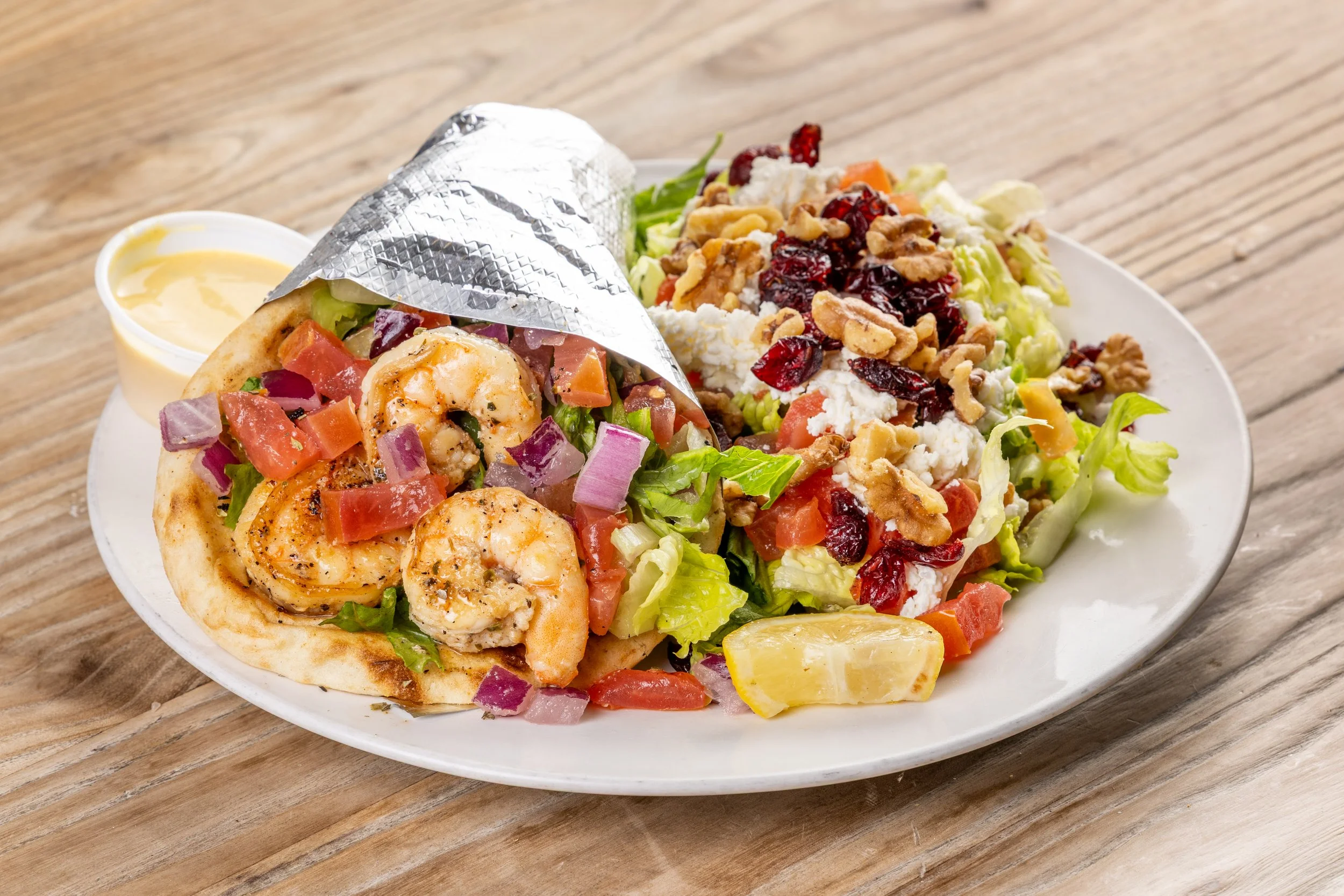 A plate with a shrimp pita, a side salad topped with nuts and cranberries, and a small cup of creamy dressing on a wooden table.