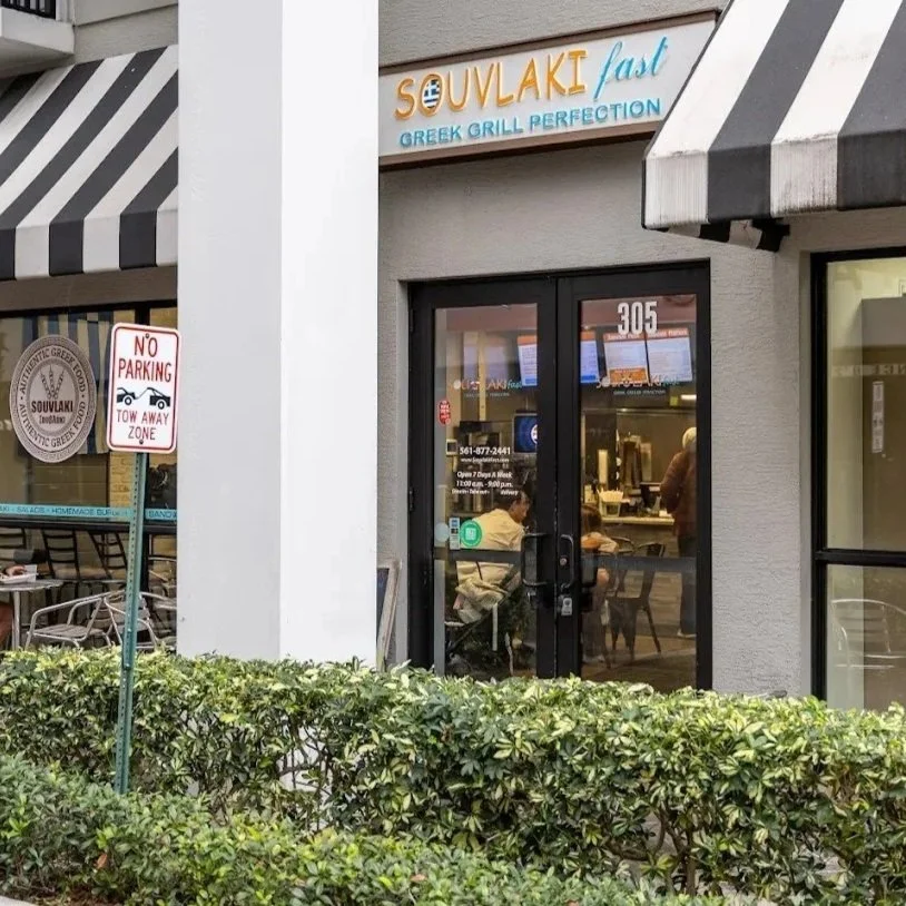 Exterior of a Greek restaurant in Boynton Beach called Souvlaki Fast with outdoor seating in between well-maintained bushes and large glass windows.