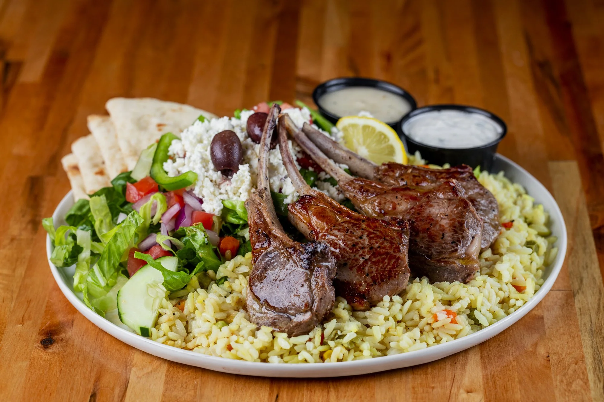 Plate of lamb chops with rice, salad, pita, lemon wedge, crumbled cheese, olives, and two small bowls of sauce on a wooden table.